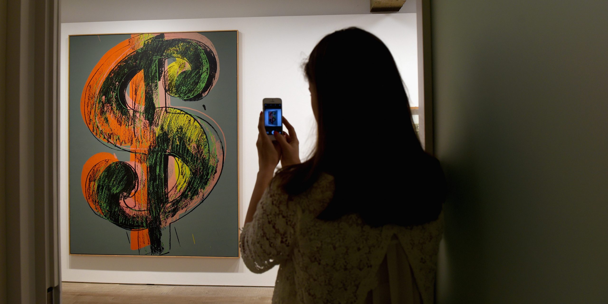 A woman takes a photograph on her mobile phone of artwork by Andy Warhol called Dollar Sign.