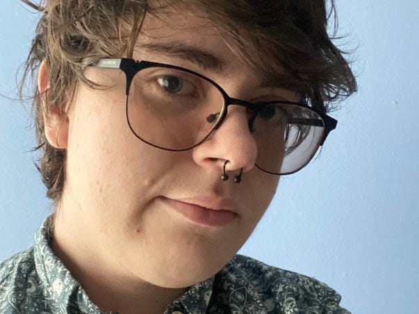 Cole Sorenson: A young, white, transmasculine person with short brown hair, tinted glasses, and a septum piercing. He is wearing a shirt with a space print.
