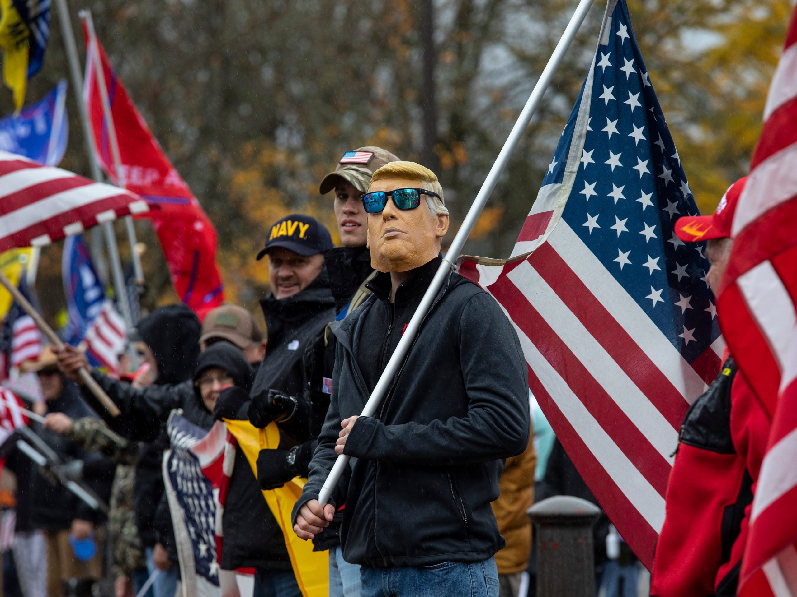 A group of Trump supporters stand carrying American flags, with one wear a Trump mask and sunglasses