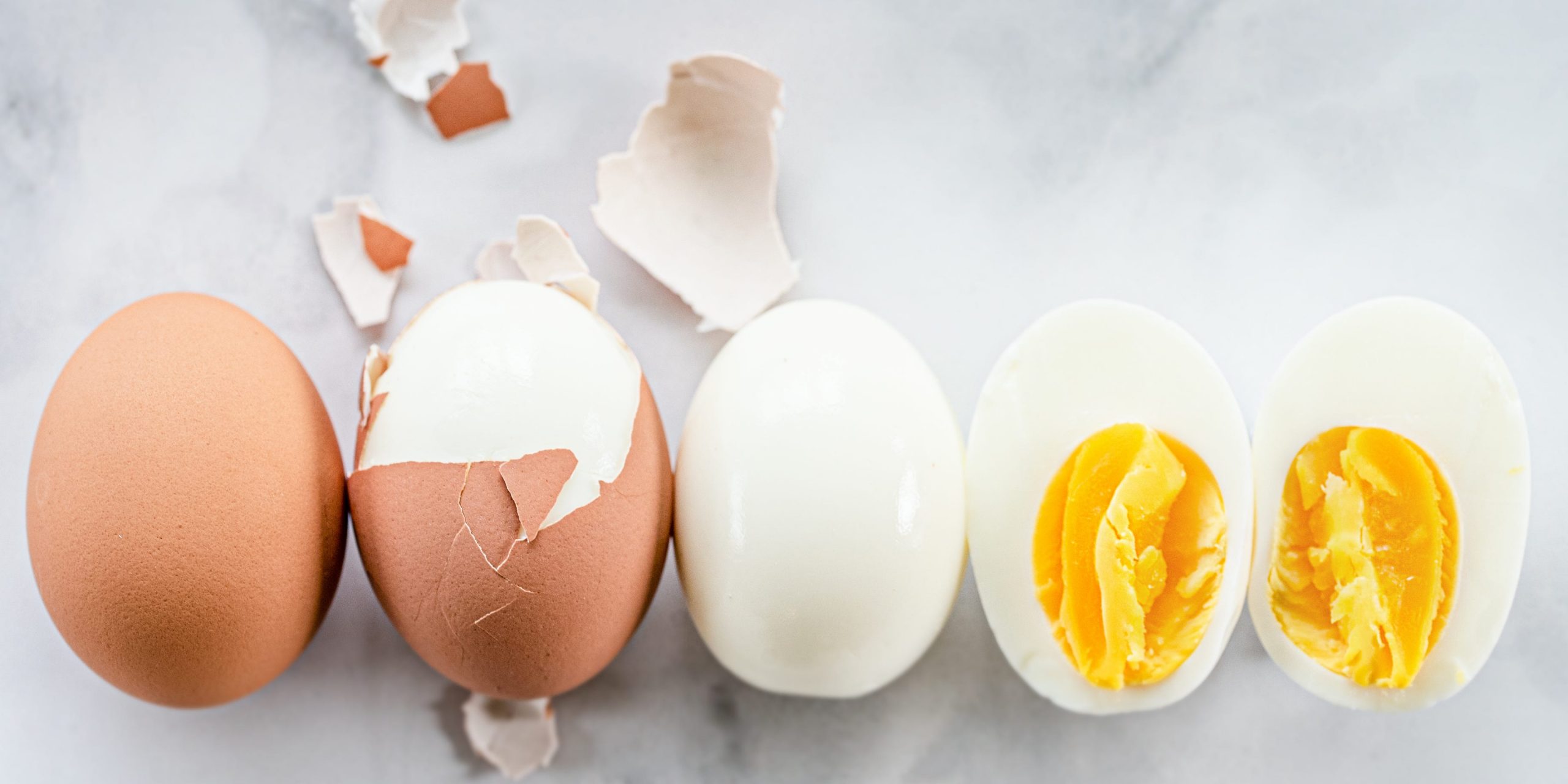 A whole egg in the shell lined up next to a partially peeled hard boiled egg, a fully peeled hard boiled egg, and a sliced open hard boiled egg