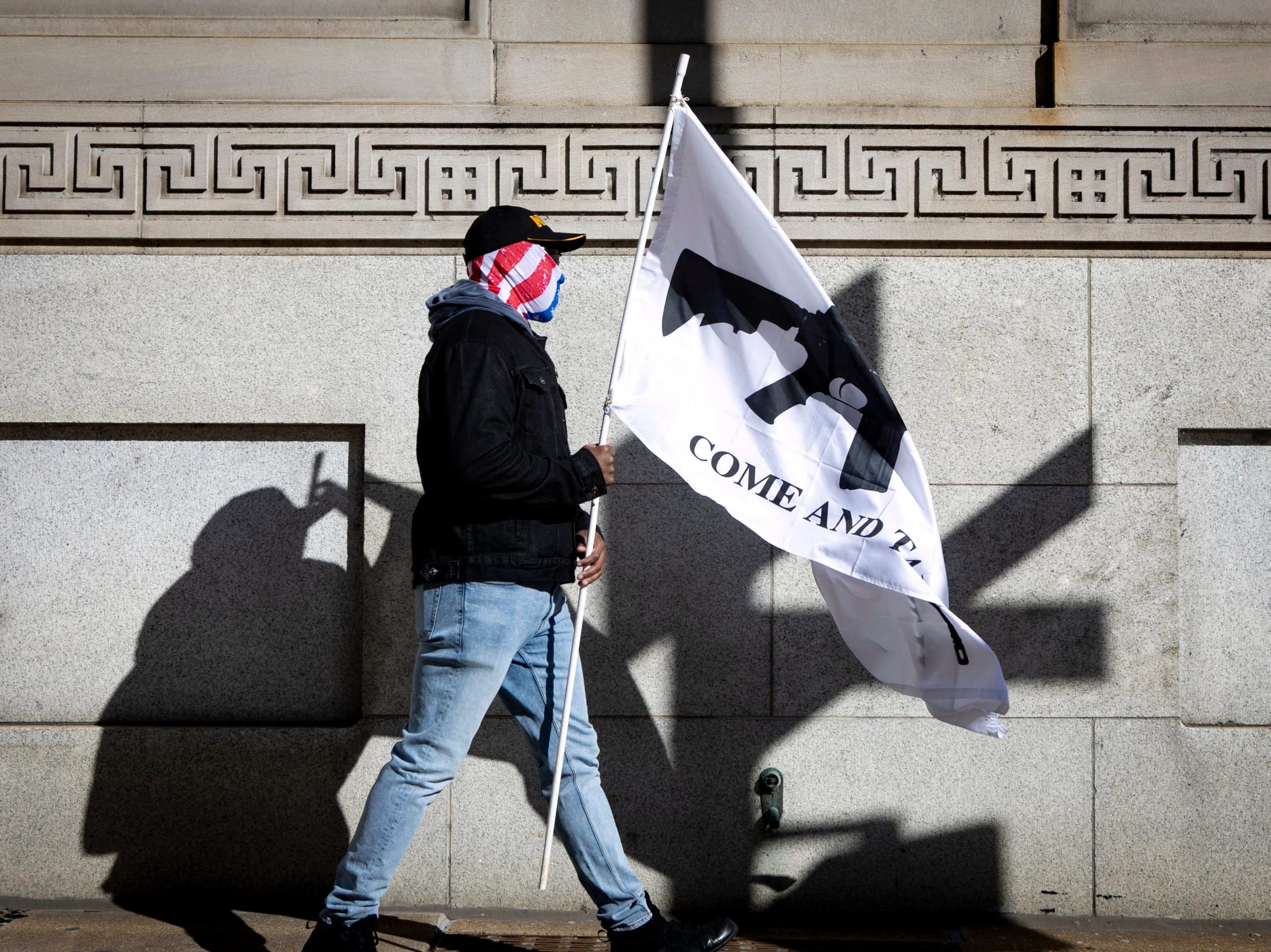 A man walks with an American flag mask over his face and carries a white flag with the image of a gun that reads "come and take it."