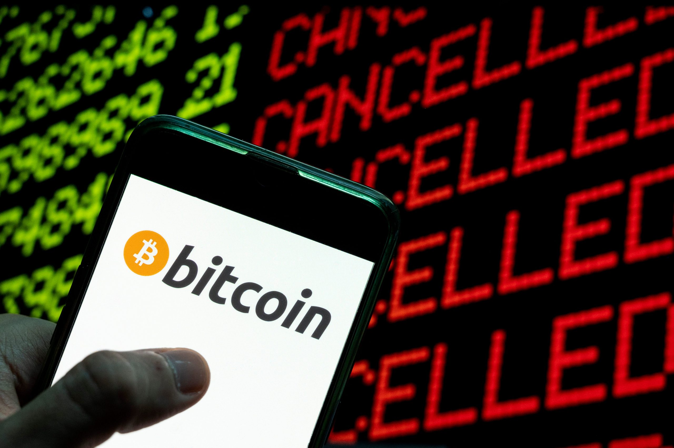 The bitcoin logo is seen on a smartphone screen device in front of a computer screen that says "cancelled. "