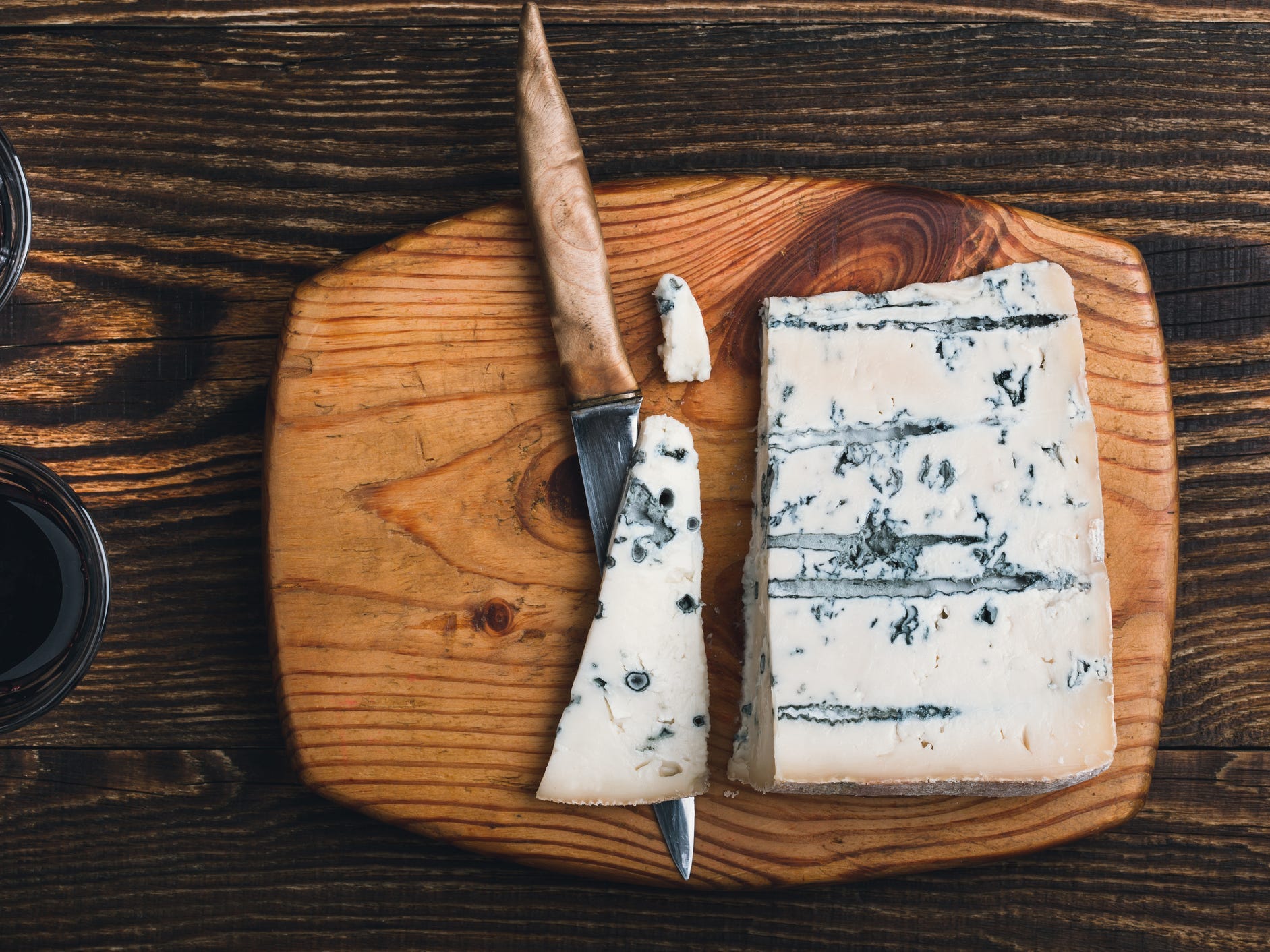 A wedge of blue cheese on a wooden cutting board with a knife and two glasses of wine