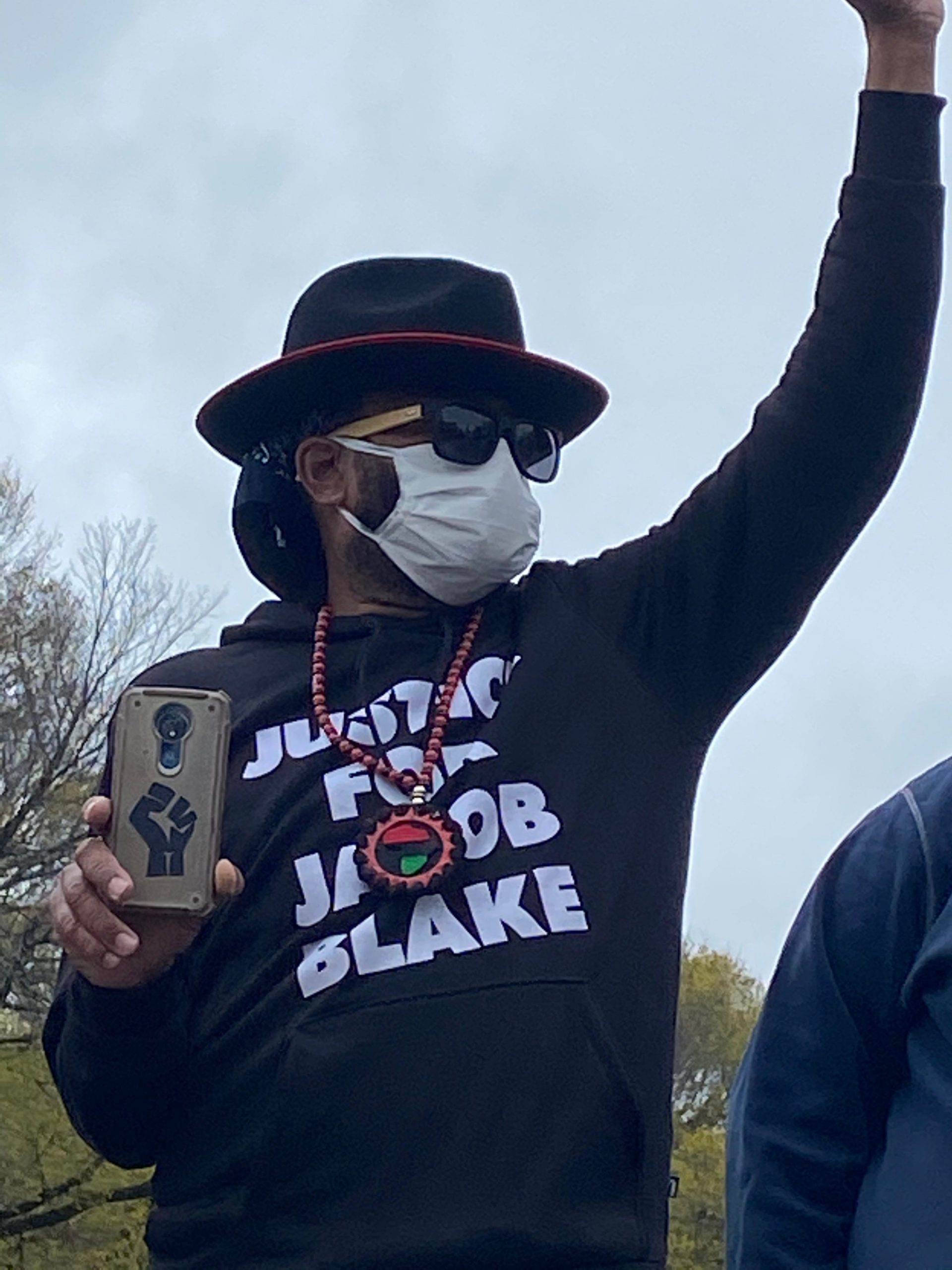 a man with a mask and a hat and a sweatshirt that says justice for jacob blake raises his fist