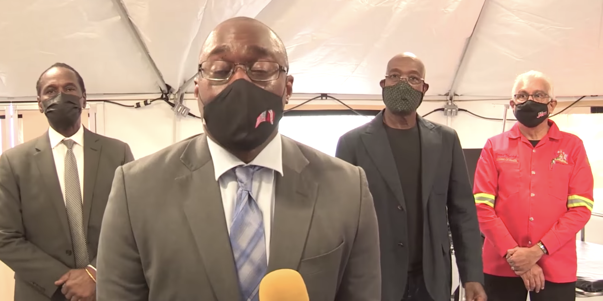 The US ambassador to Trinidad and Tobago, Joseph Mondello in a mask, speaks into a microphone in a tent with three officials behind him.