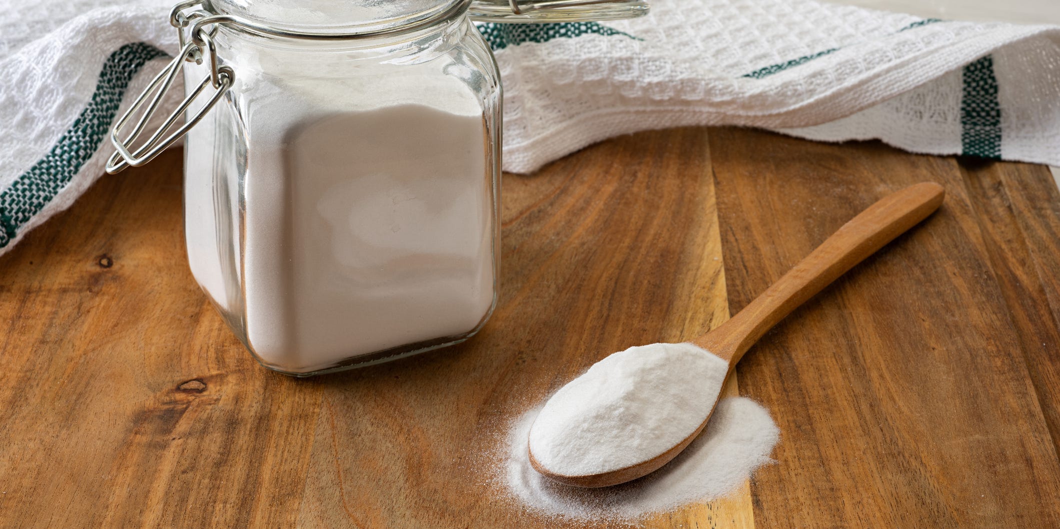 A glass jar of baking powder with a wooden spoonful of baking powder next to it on a wooden counter