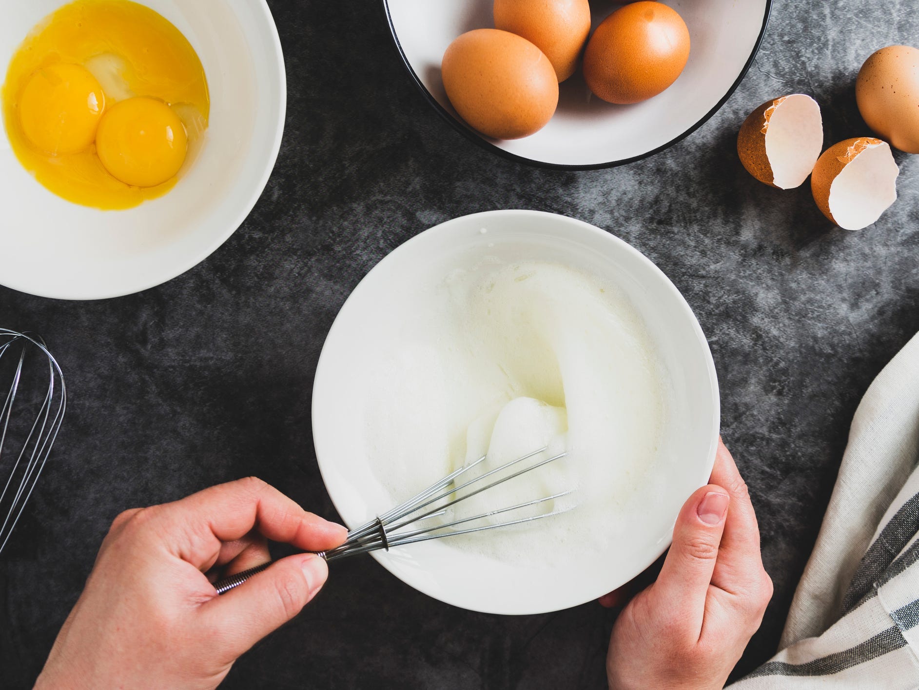 Hands whipping a bowl of egg whites surrounded by a bowl of yolks and a bowl of whole eggs