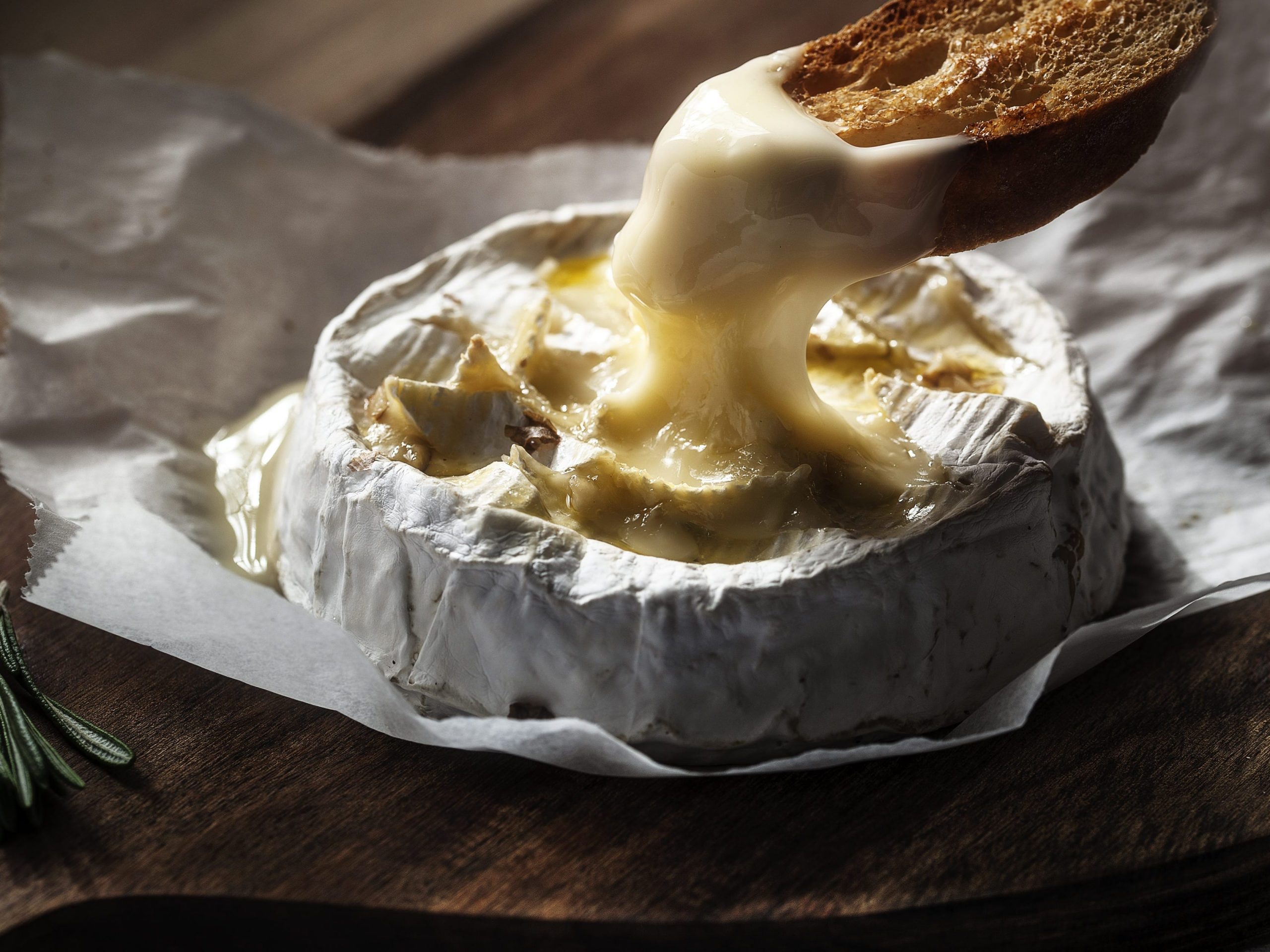 A crostini being dipped into a gooey wheel of camembert cheese