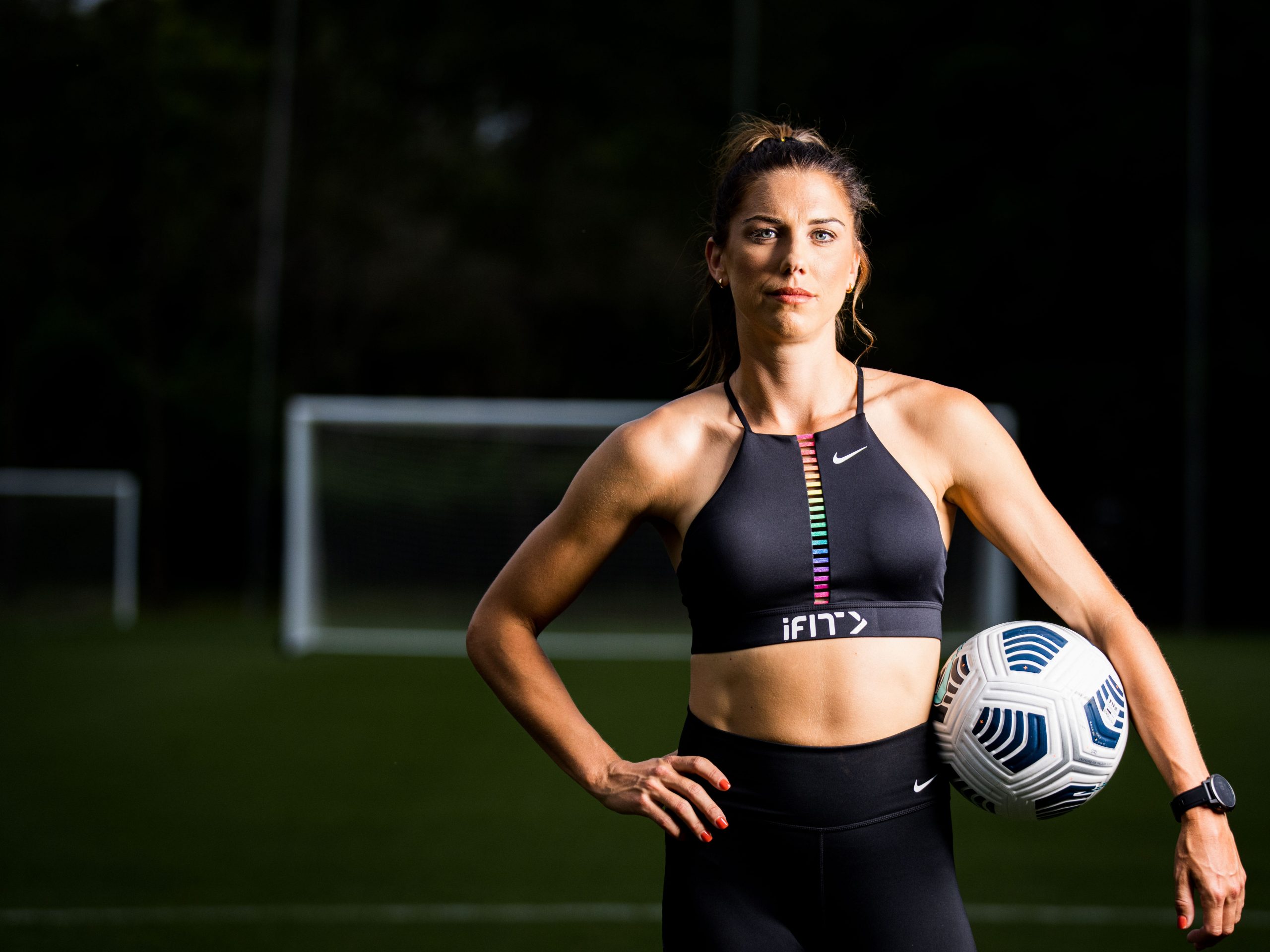 US soccer player Alex Morgan wearing iFit gym clothes and holding a soccer ball under one arm on a soccer field.