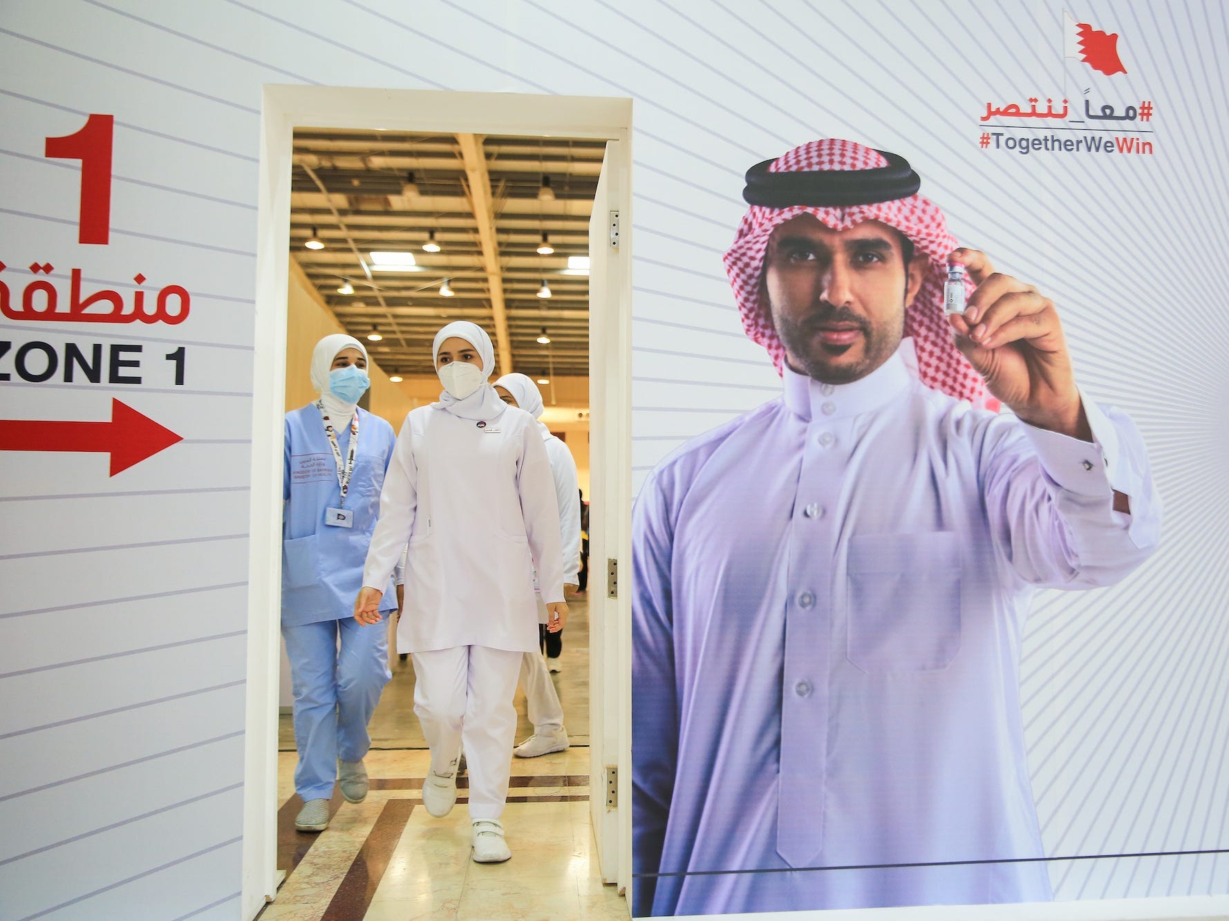 Nurses wearing masks walk towards a doorway, on the wall, a poster or a man is holding a vial of COVID-19 vaccine.