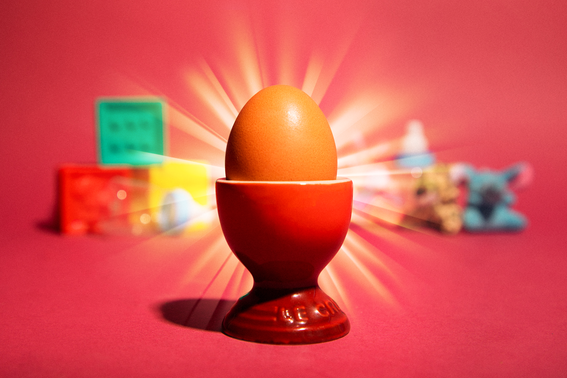 A photo of a radiating egg in an egg cup with blurred children's toys behind it on a red background.