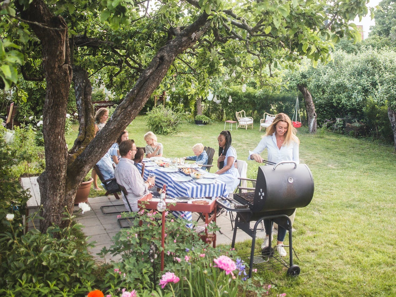 People sit at a table in a backyard while someone grills next to them.