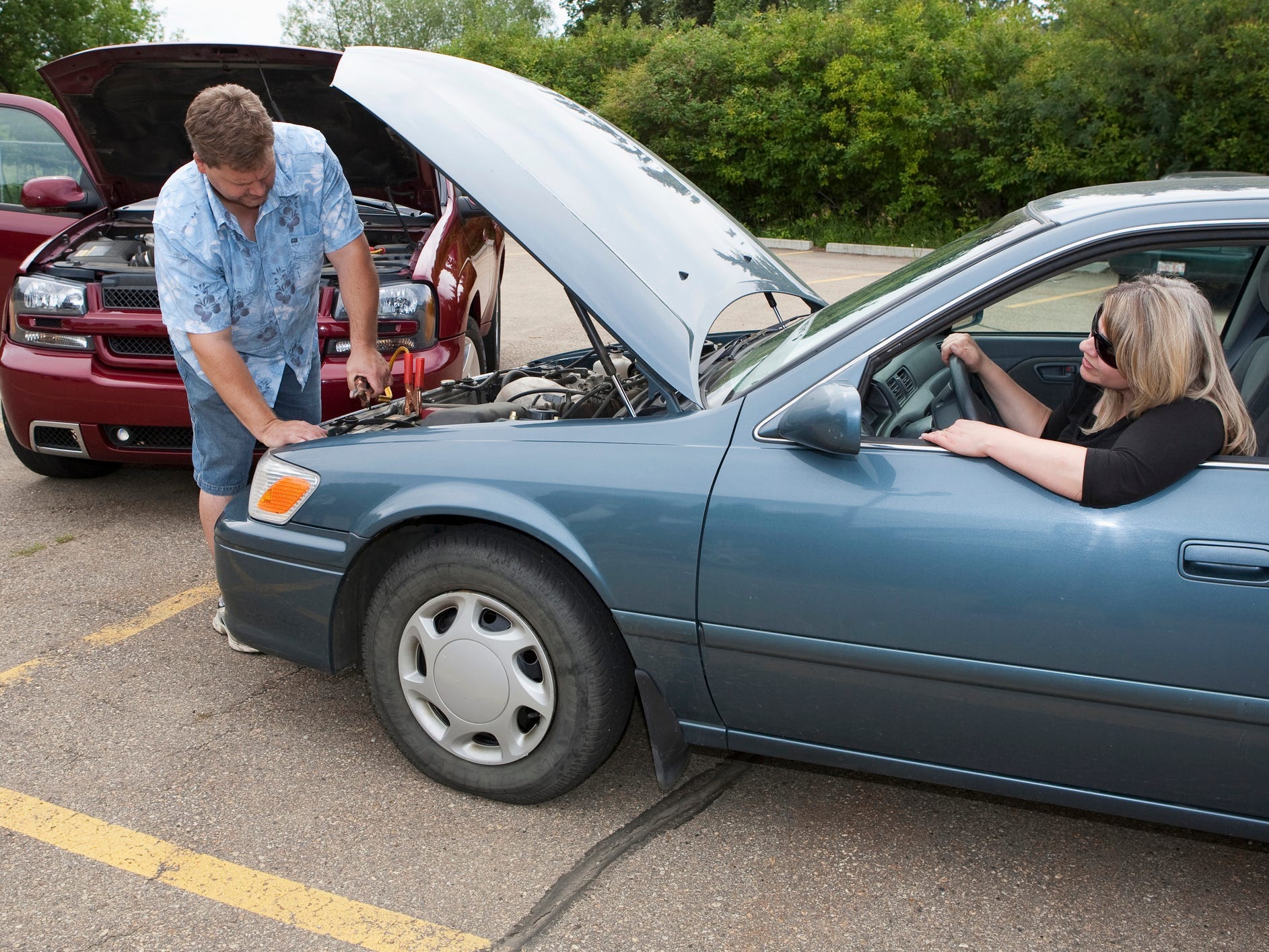 A person hooks up jumper cables to the battery of a blue car while the driver looks on