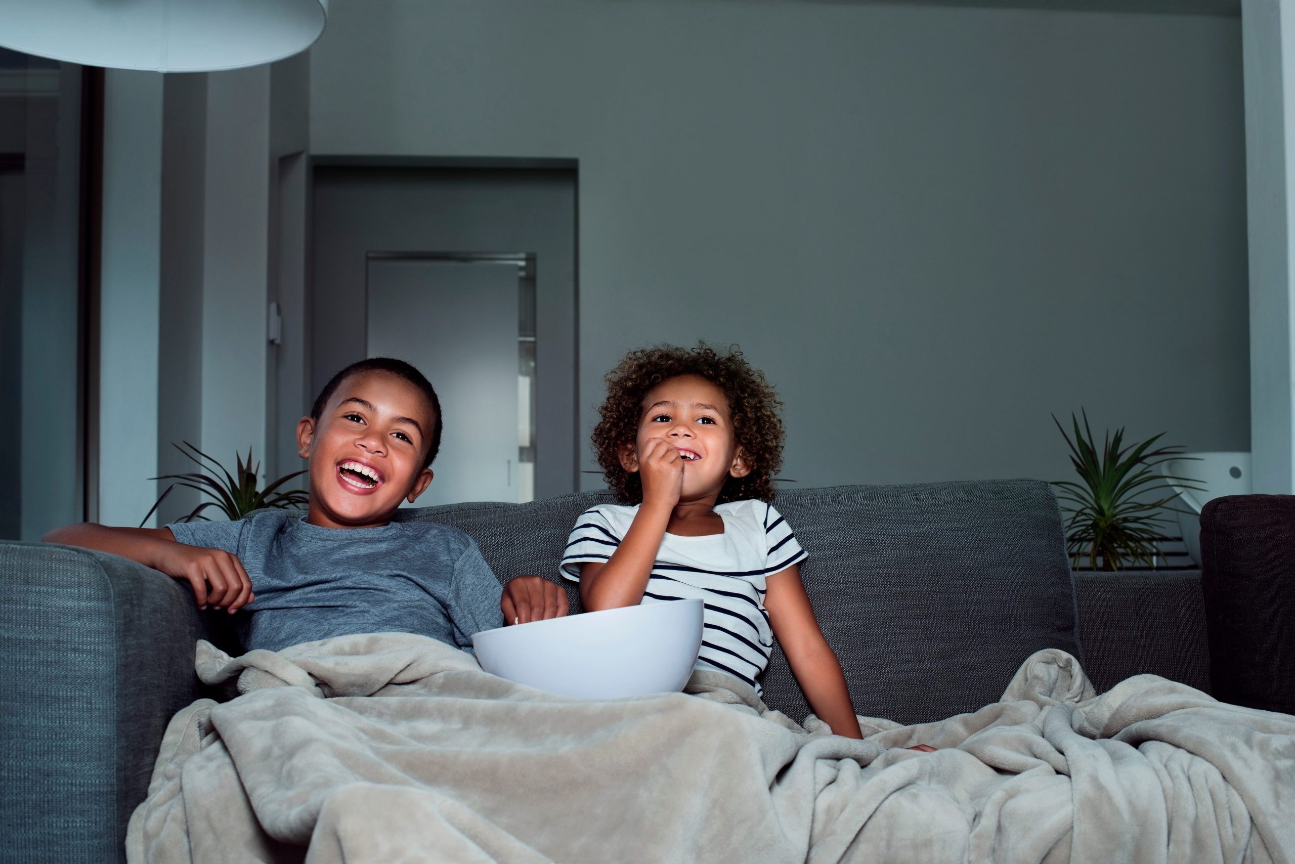 Two children sitting on a couch watching TV and eating popcorn