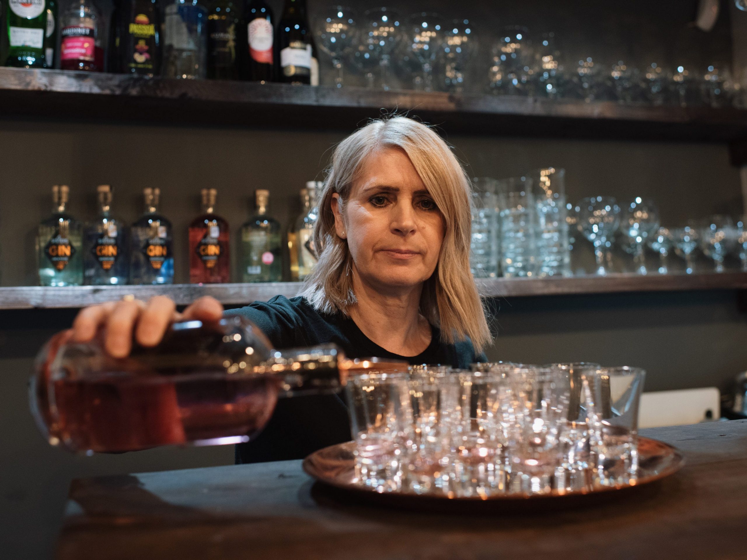 A woman behind a bar pours gin into shot glasses on a tray with shelves of bottles and glasses in the background