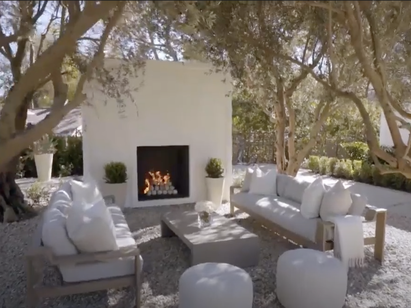 An outdoor fireplace with two couches, a coffee table, and two cushions surrounded by olive trees.