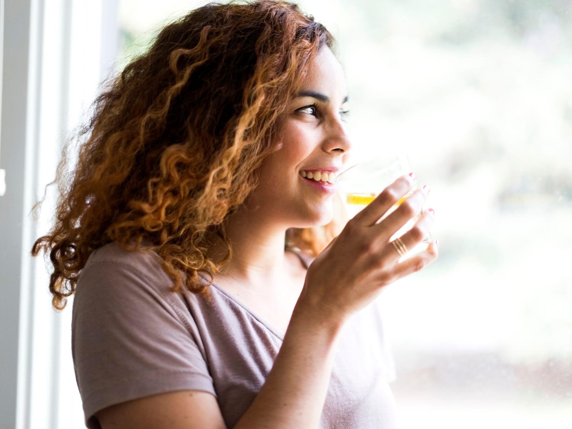 A person with shoulder-length curly hair drinking a glass of water with lemon in it next to a window view of the outdoors.