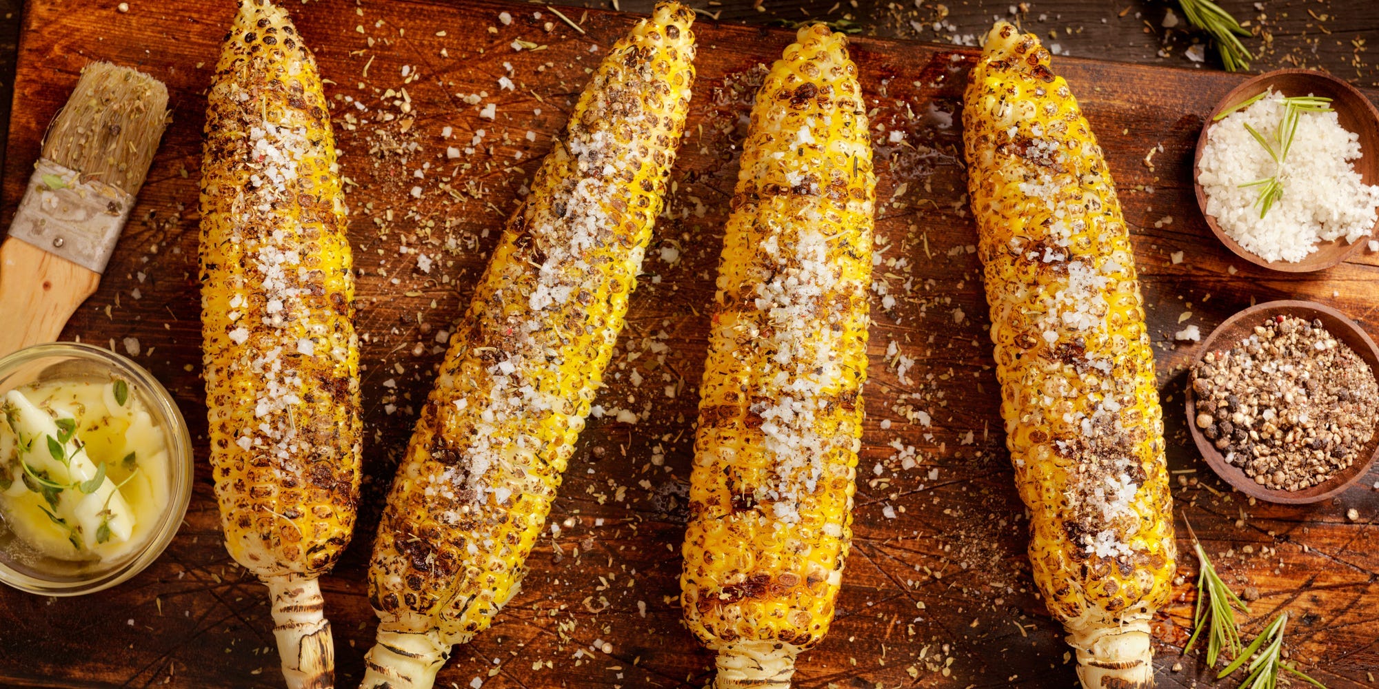 Four ears of grilled corn with char marks seasoned with salt and pepper sitting on a wooden cutting board