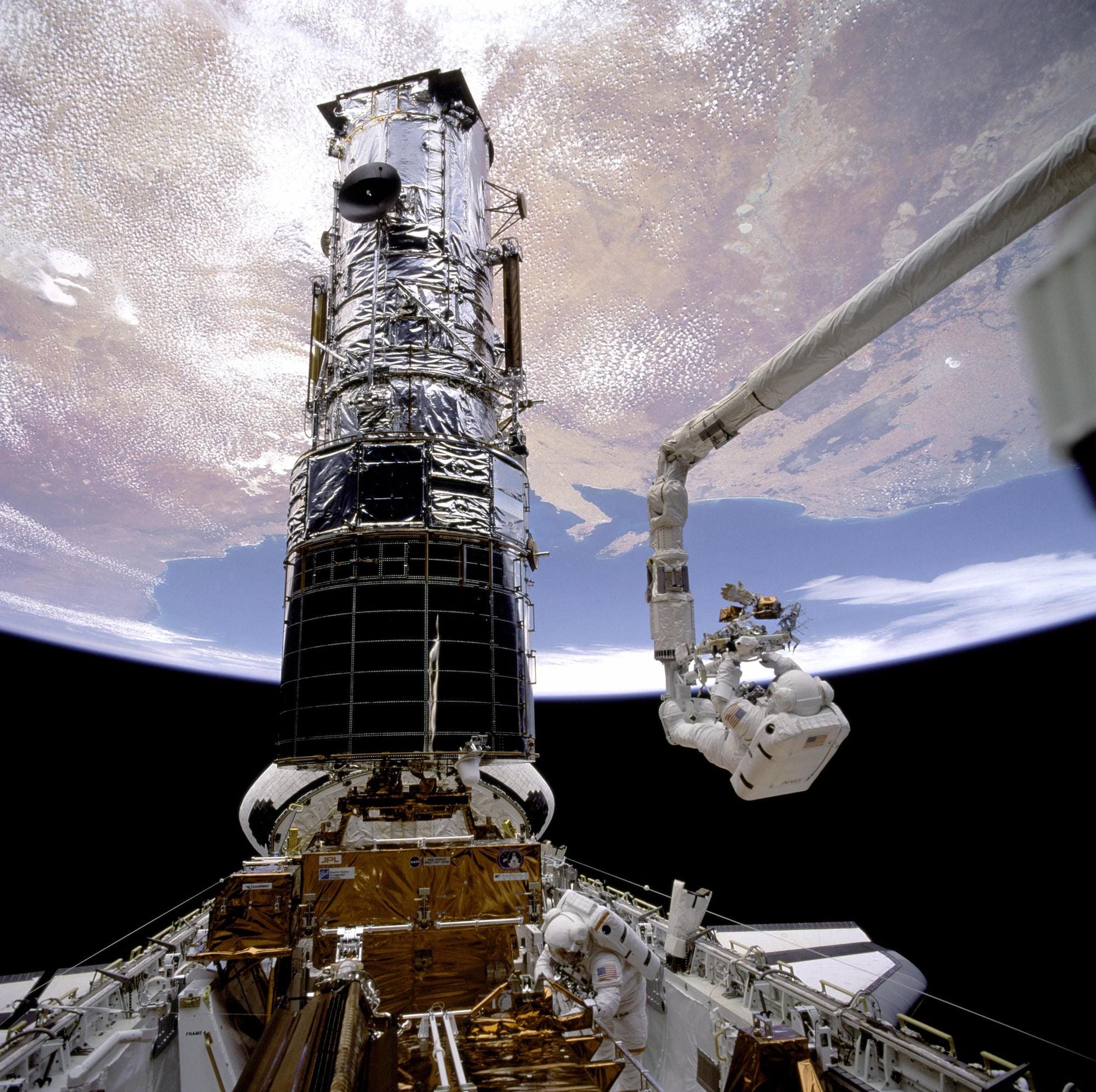 two astronauts in spacesuits work on the hubble space telescope in space above earth