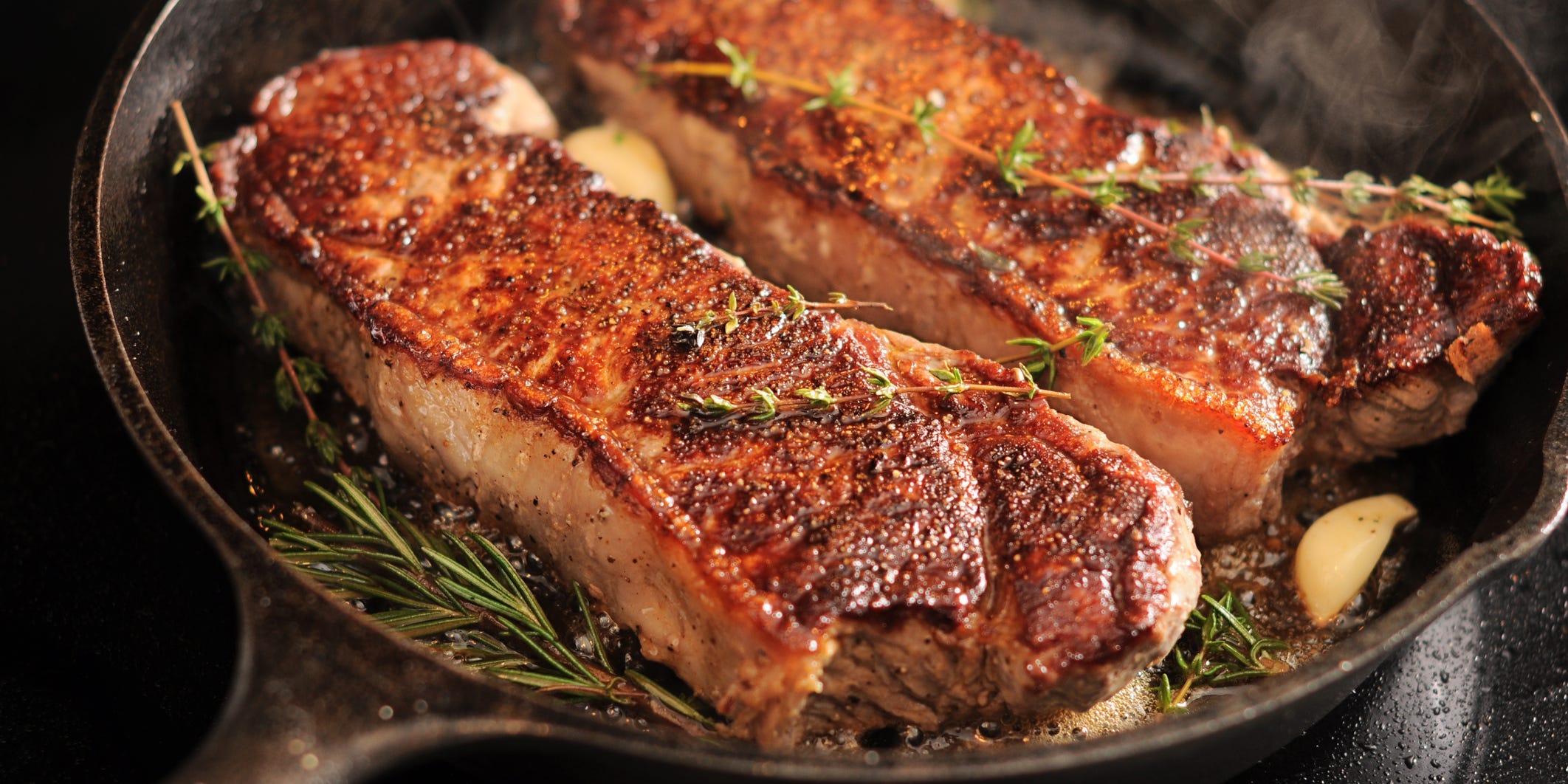 Two steaks in a cast iron pan seared to golden brown topped with whole garlic cloves and thyme