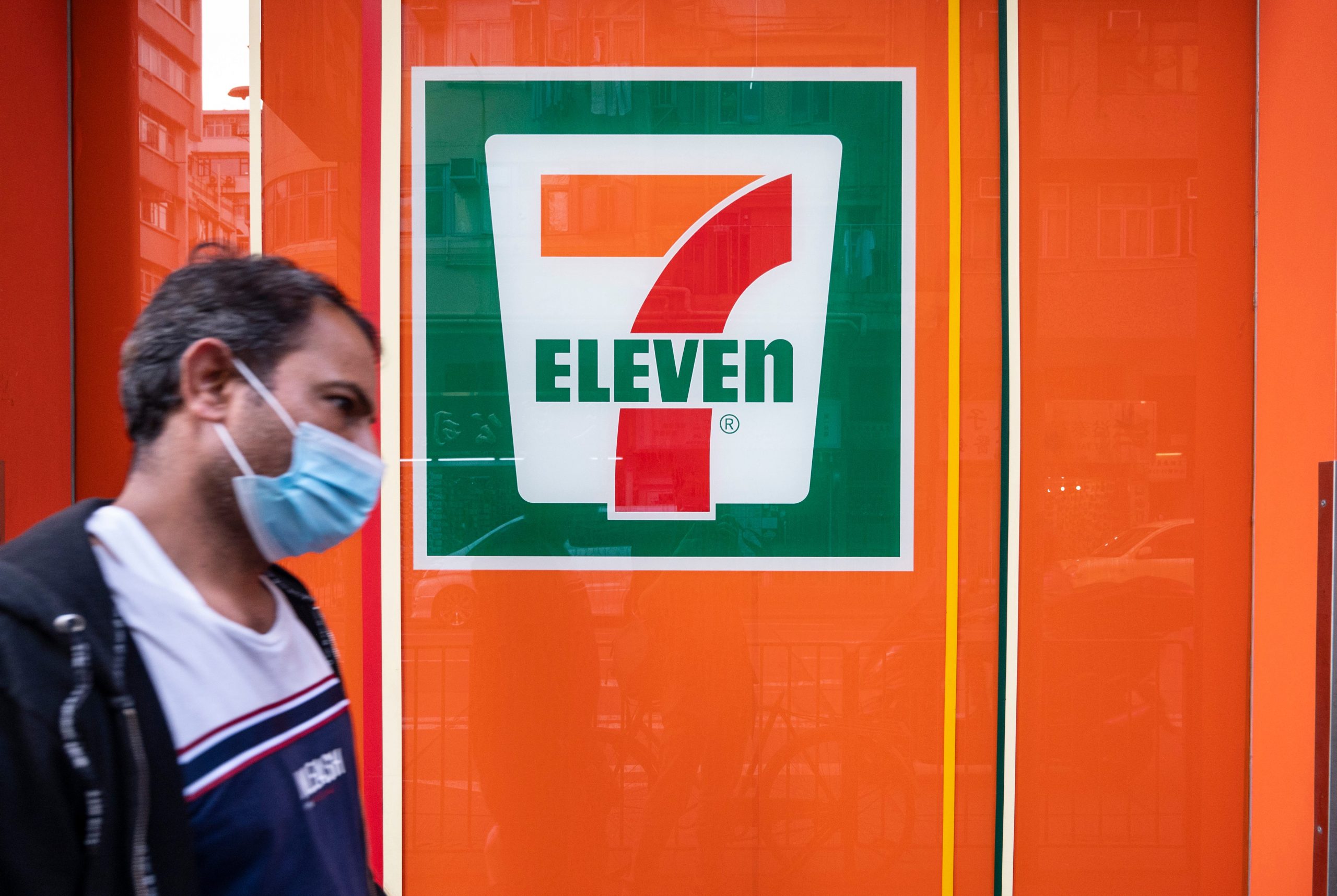 A man wearing a facemask walks past a 7-Eleven sign on an orange wall