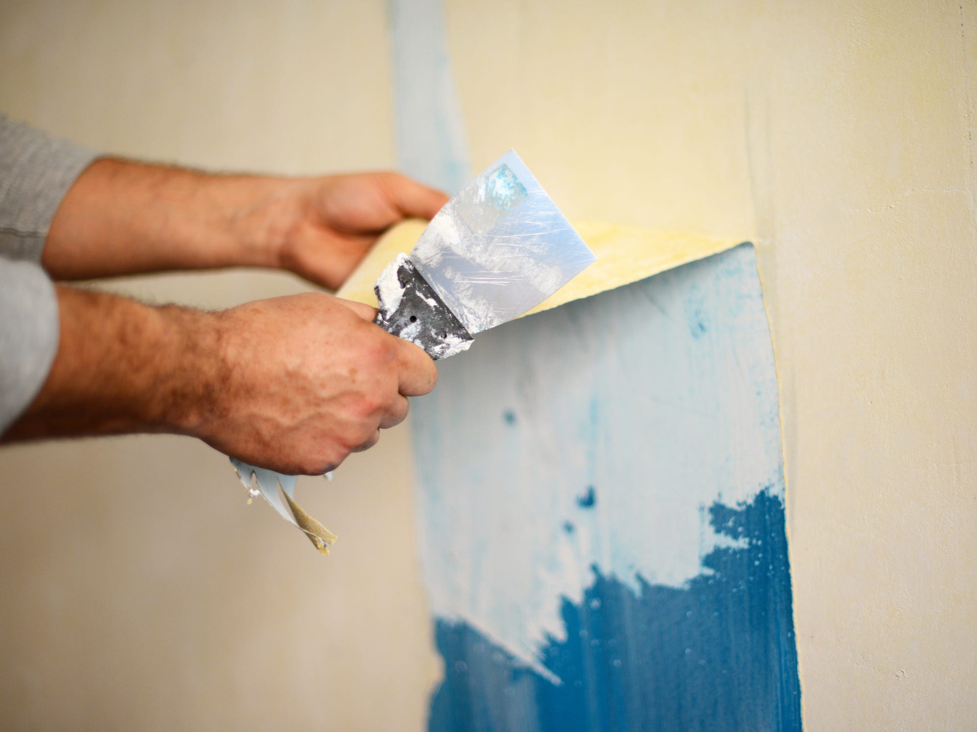 A pair of hands holding onto a strip of wallpaper and a putty knife and pulling the wallpaper off of the wall