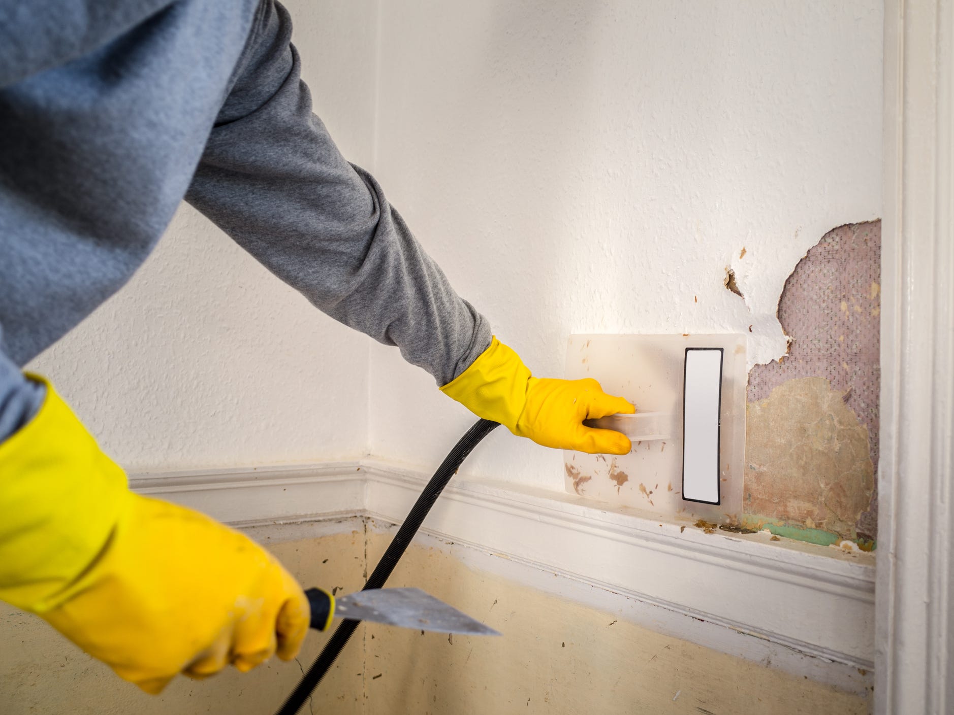 A person wearing yellow rubber gloves applying a wallpaper steamer to wallpaper