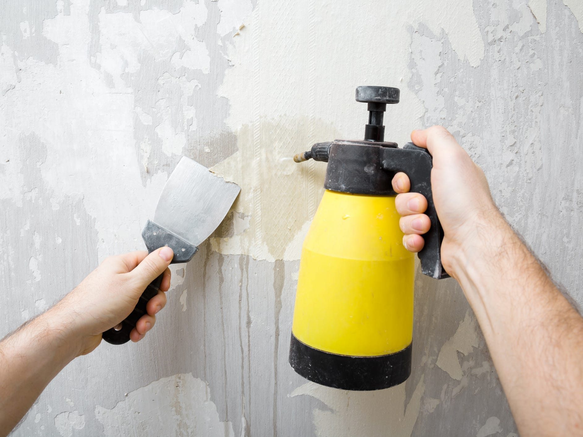 One hand holding a putty knife and another hand spraying solvent onto a wall out of a yellow spray bottle