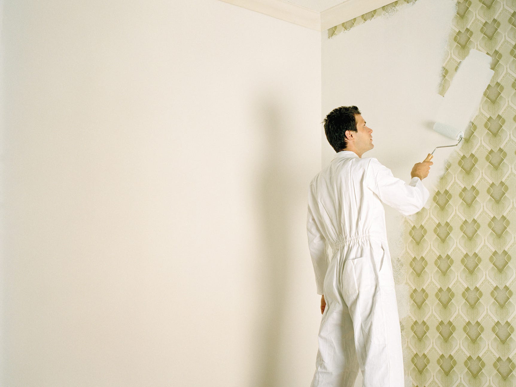 A person wearing white coveralls paints over patterned wallpaper