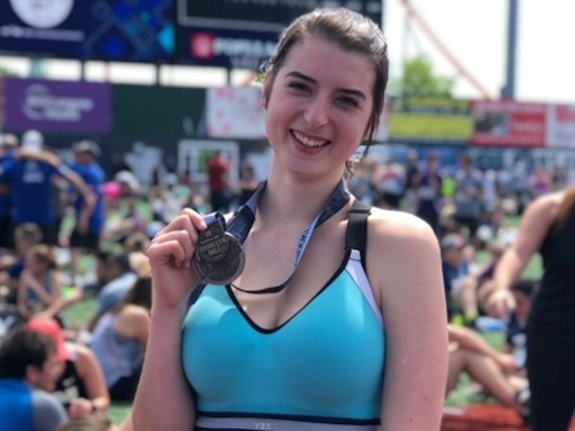 Rachel Robles, wearing a blue tank top, holds up a medal from Popular Brooklyn Half Marathon she ran in 2019. In the background are other runners sitting on the ground.