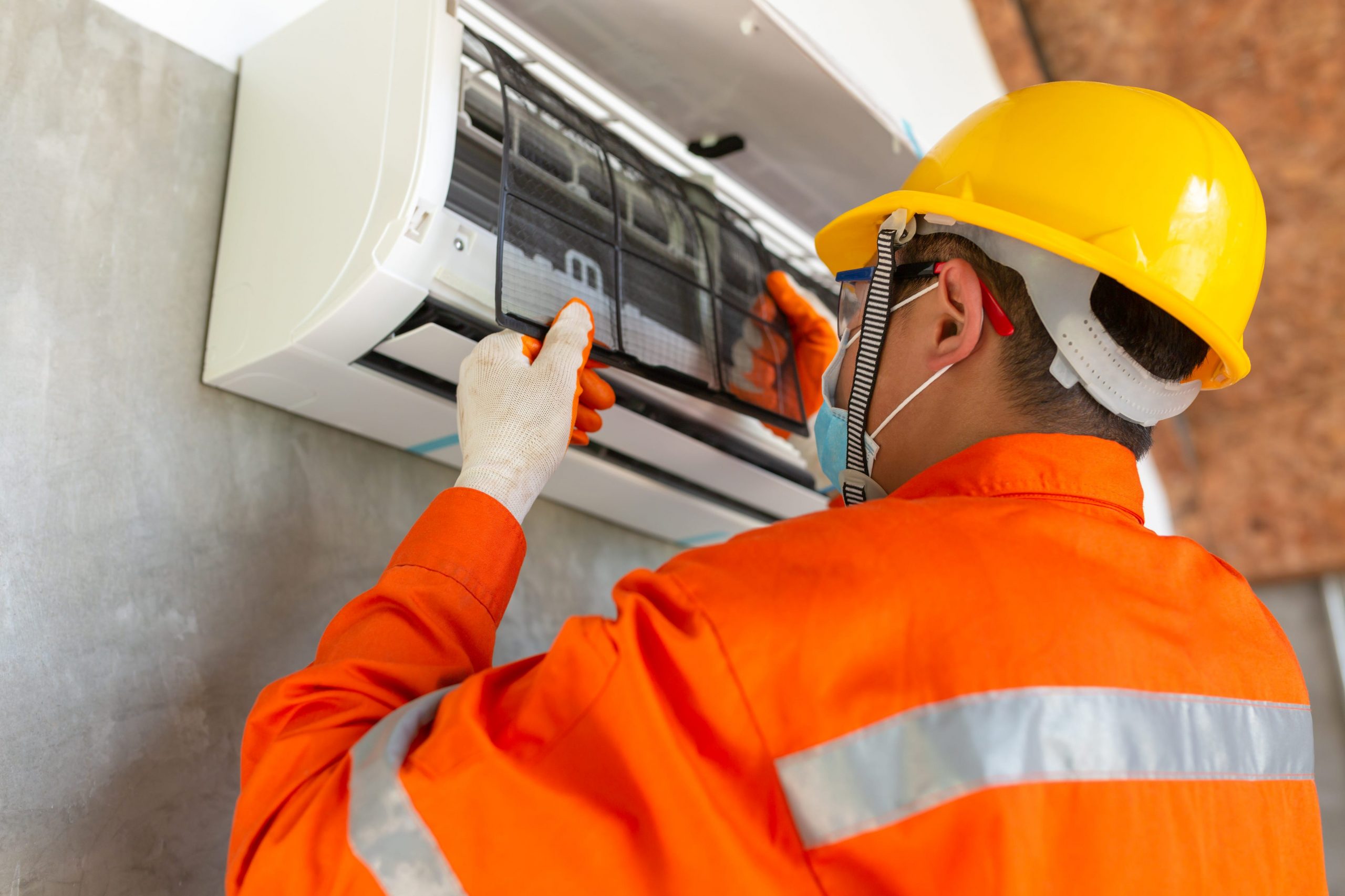 A technician instals an air conditioning unit in a house wearing a bright orange jacket.
