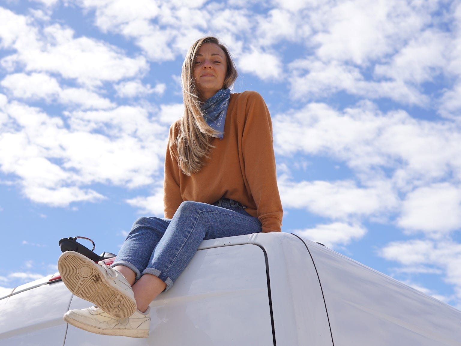 A woman wearing an orange sweater sits on top of a white van with the blue sky and clouds behind her.