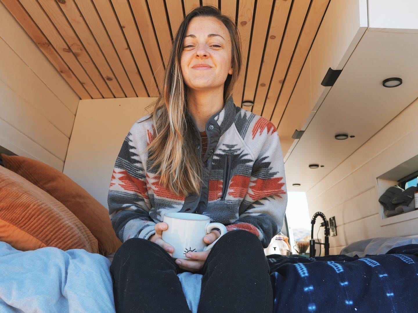 A woman wearing a grey and red sweater sits on her bed smiling and holding a mug.