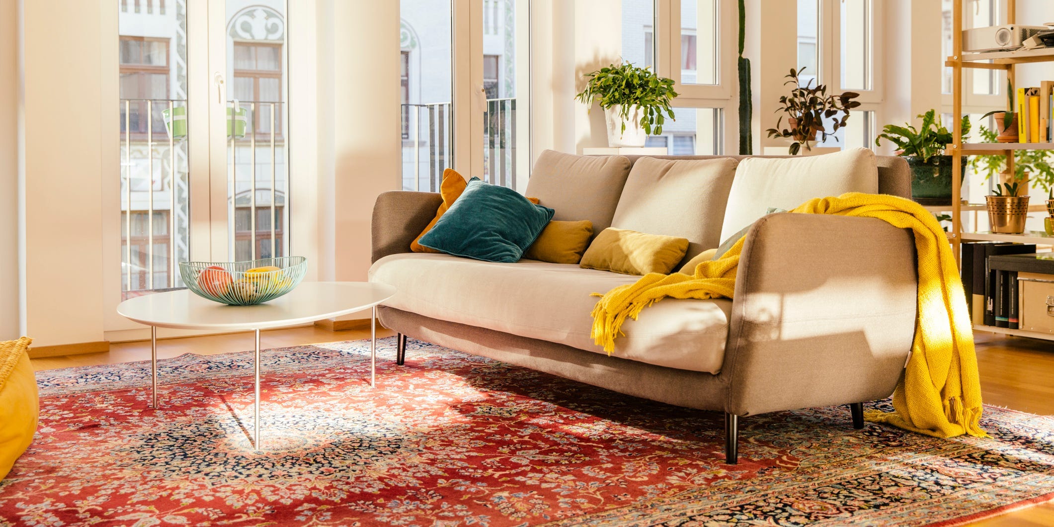 A living room with tall windows, white walls, a beige couch, a wooden coffee table, and a Persian rug