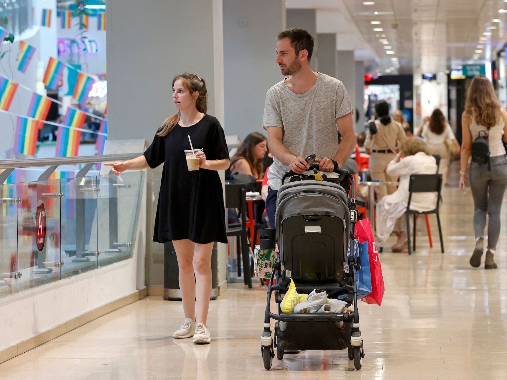 A woman and a man are pushing a stroller, maskless, in a shopping center in Tel Aviv, Israel. In the background are pride flags