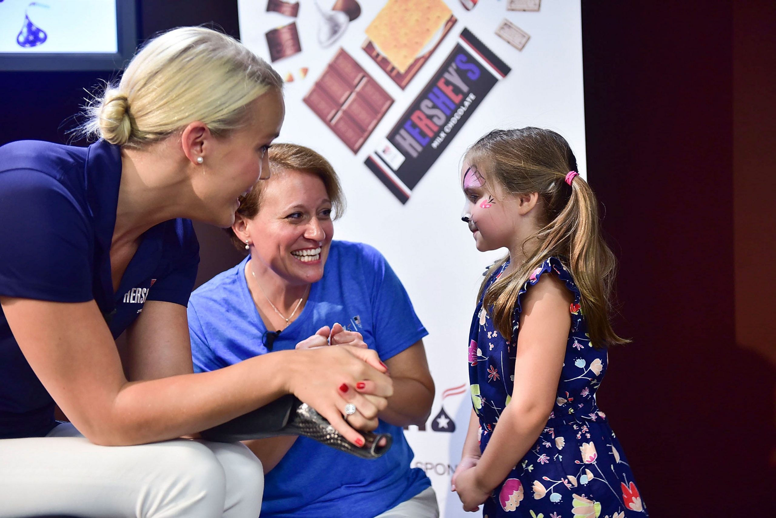 Mallory Weggemann laughs with a young girl who's a fan. Mallory is wearing a blue short-sleeve shirt with white pants, and the little girl is wearing a floral blue dress.