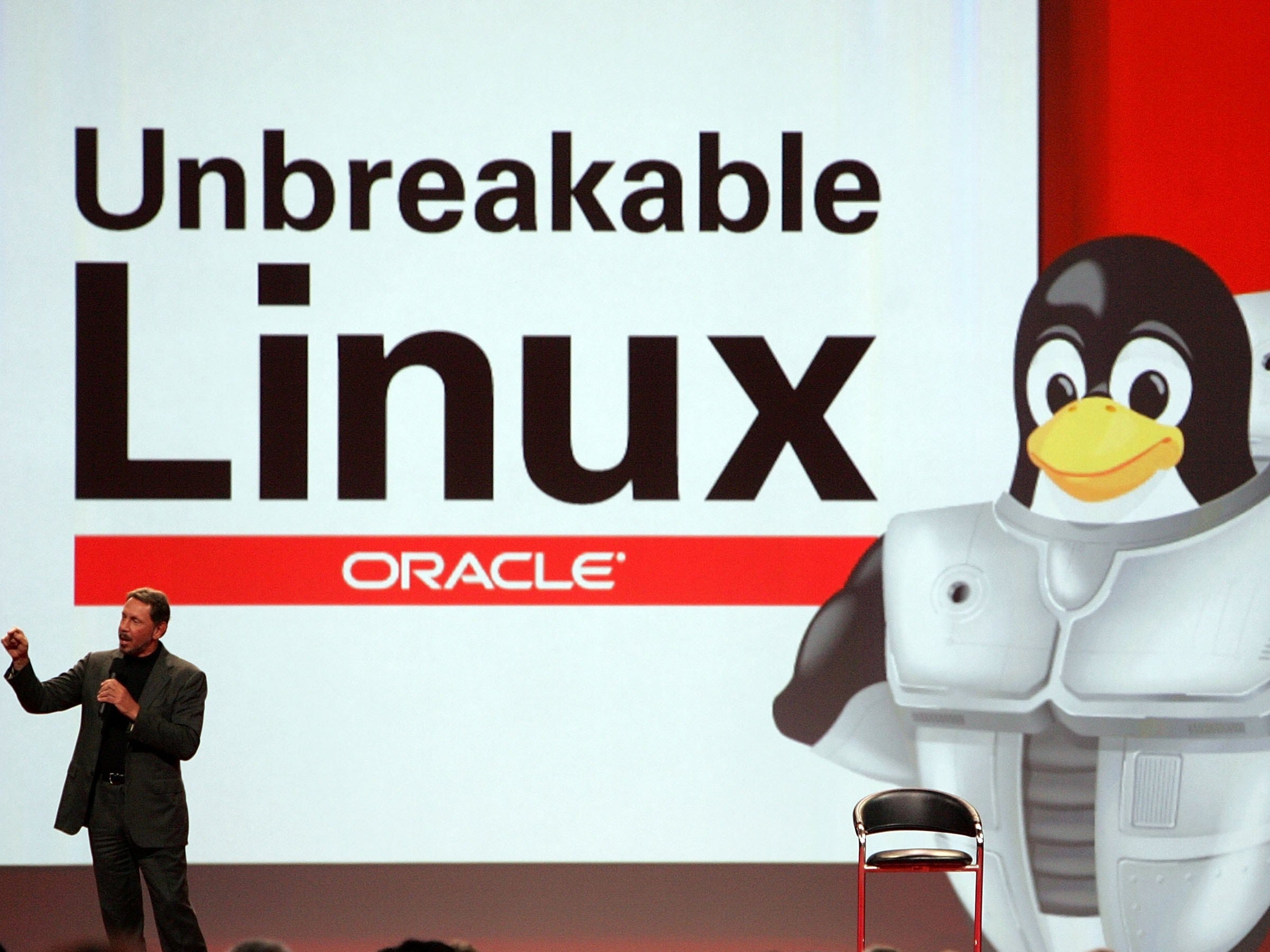 Oracle CEO Larry Ellison speaking on stage at the conference with a giant Linux sign behind him and pictures of penguins wearing armor