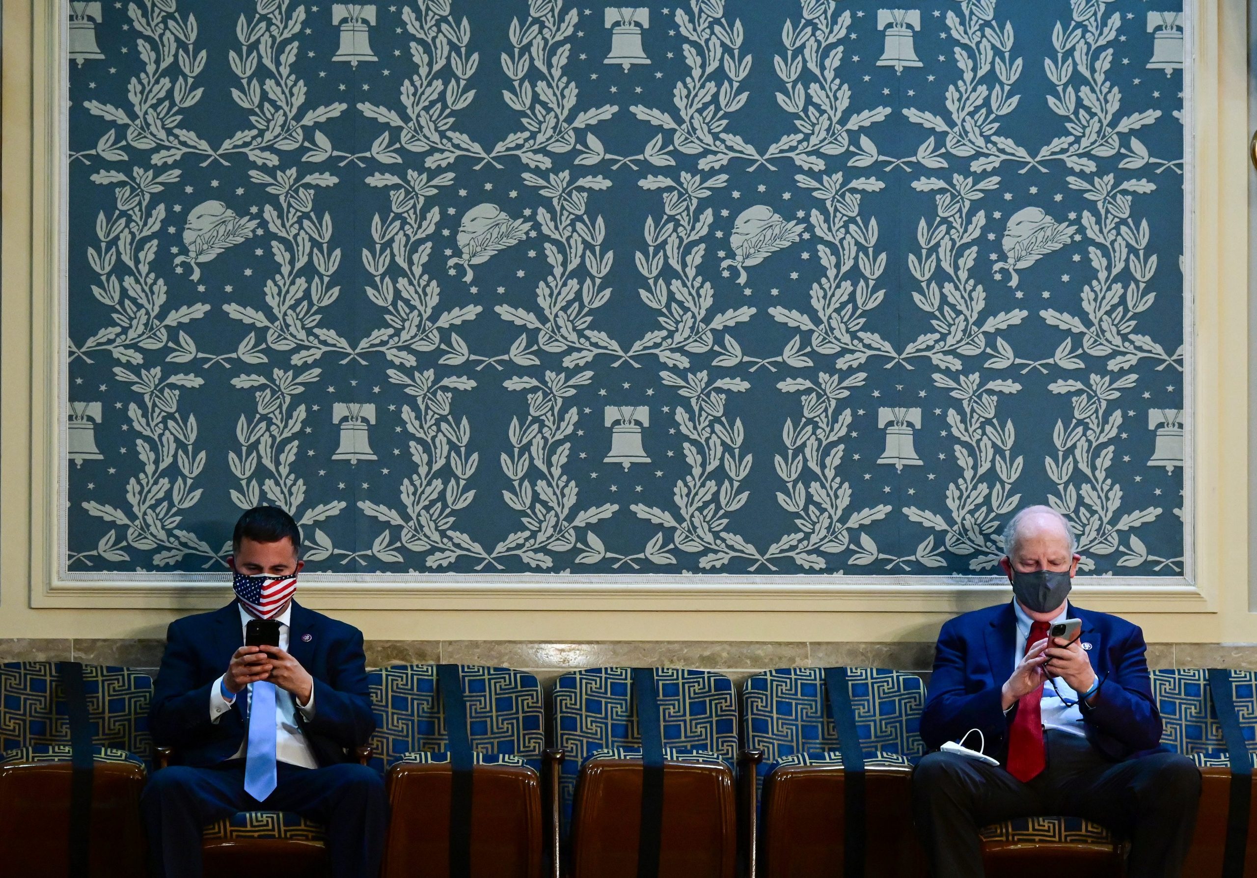 Two masked congressmen sit in front of a wallpapered wall while checking their cellphones
