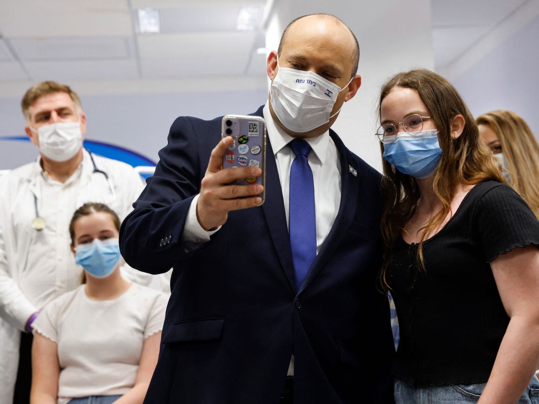 Israel's Prime minister Naftali Bennet is standing next to an adolescent, holding out a phone as he records a video message in a hospital ward. Behind him are two health workers and a child looking on