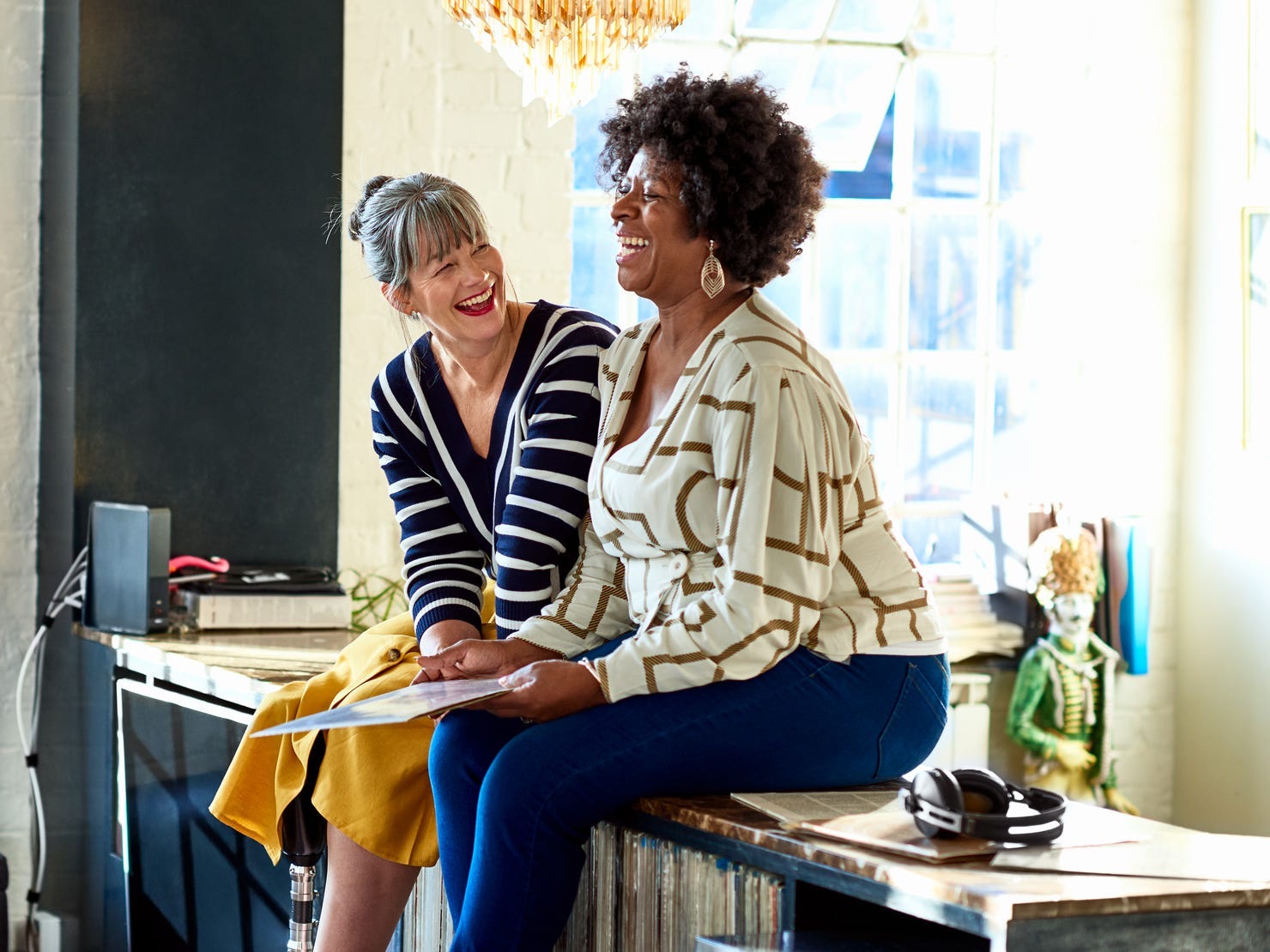 Mature women laughing together in stylish loft apartment. Two female friends sitting on sideboard and smiling.