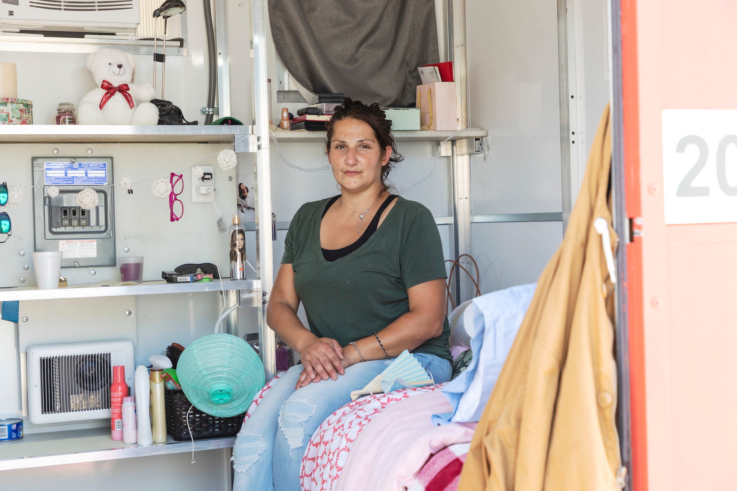 A woman sits on the bed with a shelf full of belongings behind her.