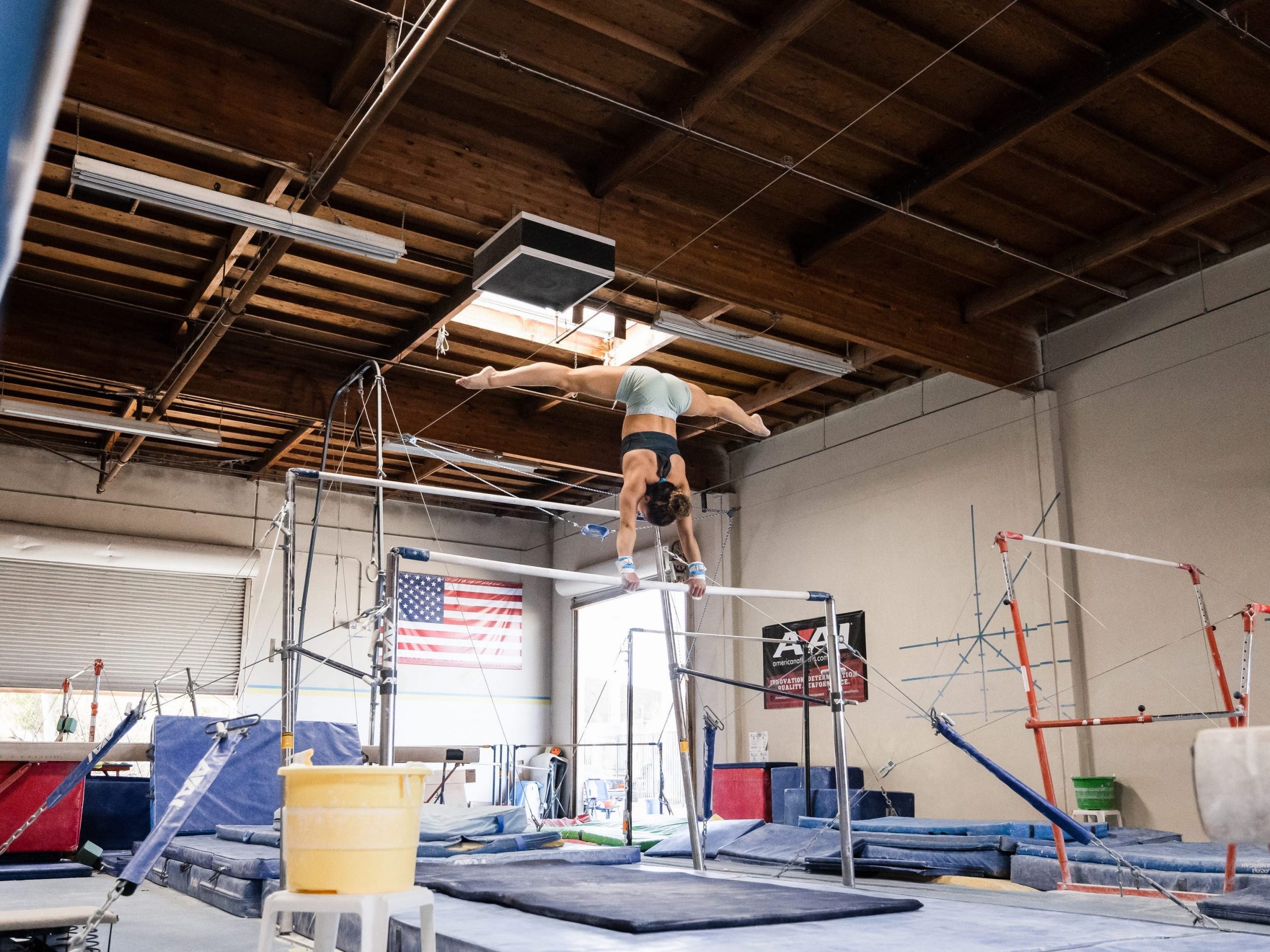 Laurie Hernandez doing a handstand splits on the uneven bars in a gymnastics gym.