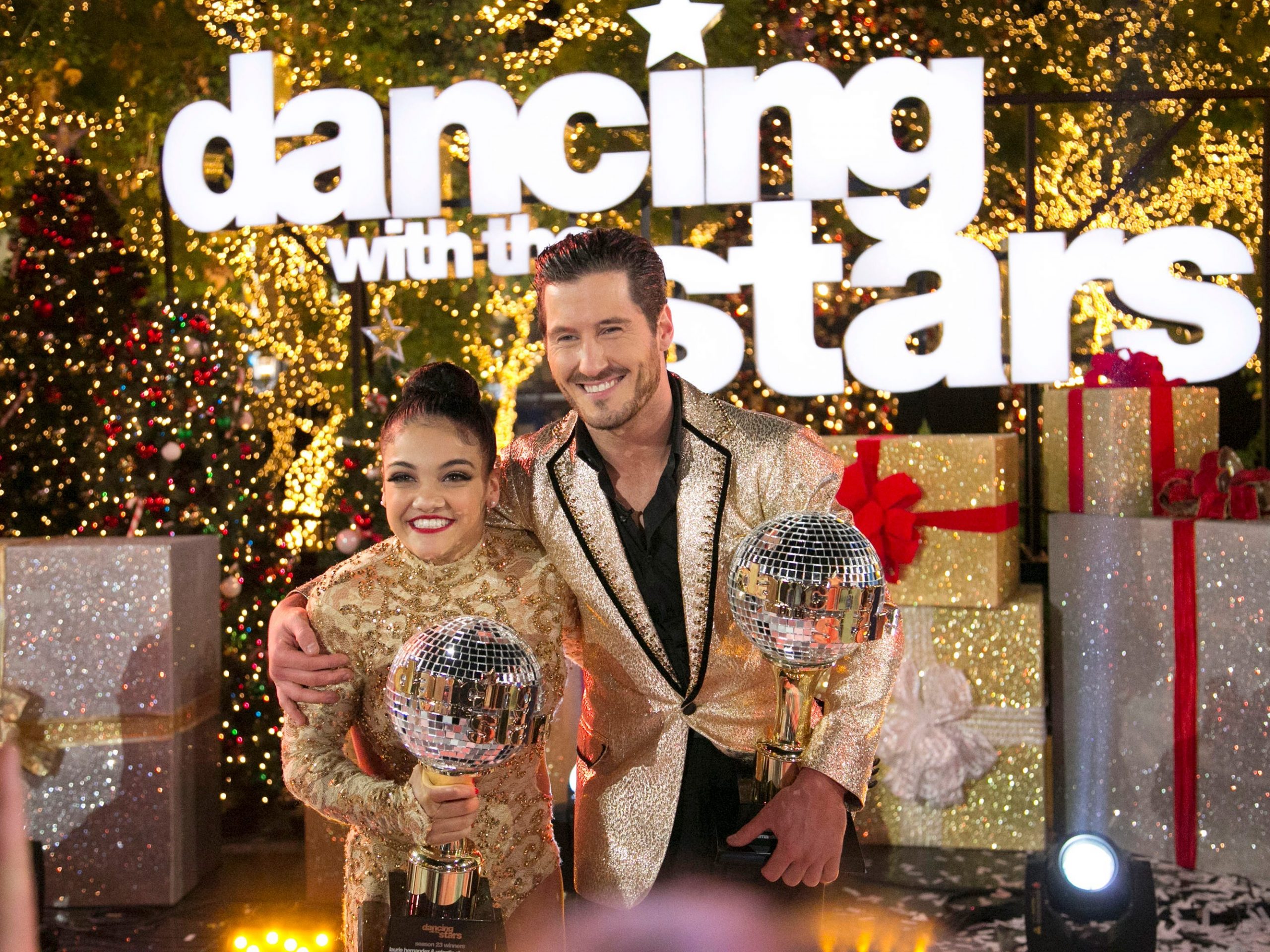 Laurie Hernandez with her trophy and her partner, Valentin Chmerkovskiy, with his trophy in front of a gold background that says "Dancing with the Stars."