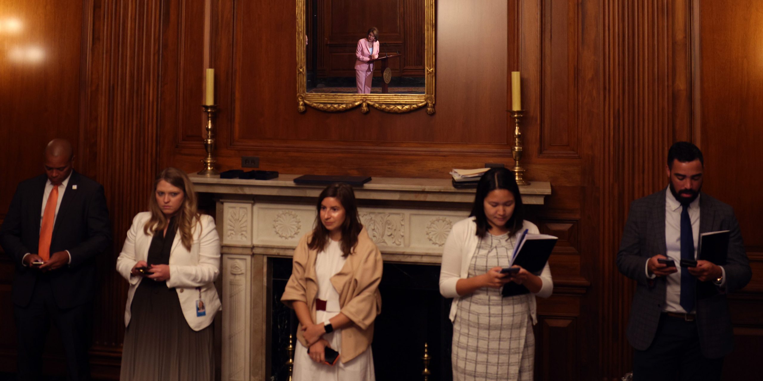 Capitol Hill staffers standing in front of an ornate fireplace listen as House Speaker Nancy Pelosi speaks