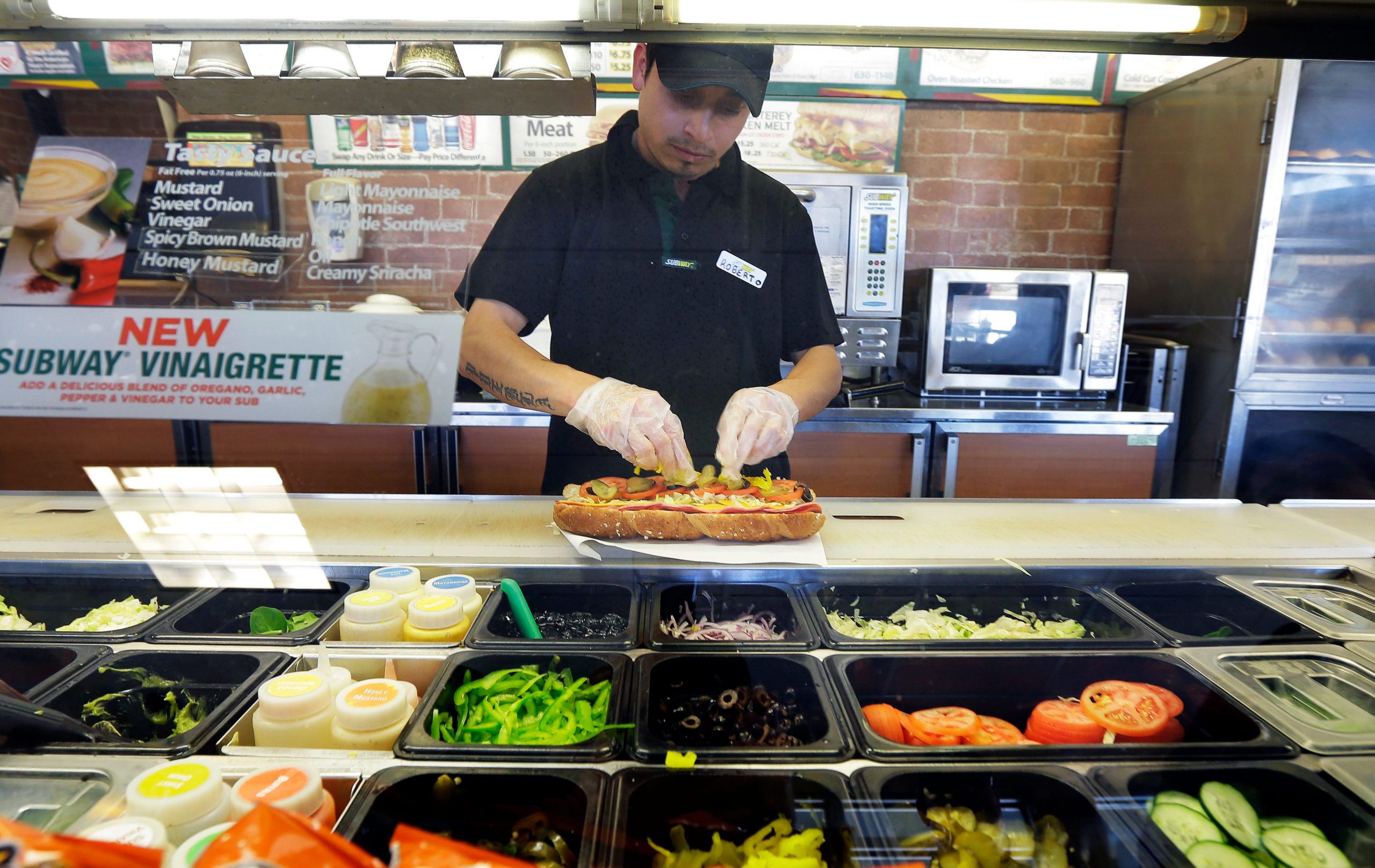 A man in a restaurant uniform makes a Subway sandwich with pickles and tomatoes