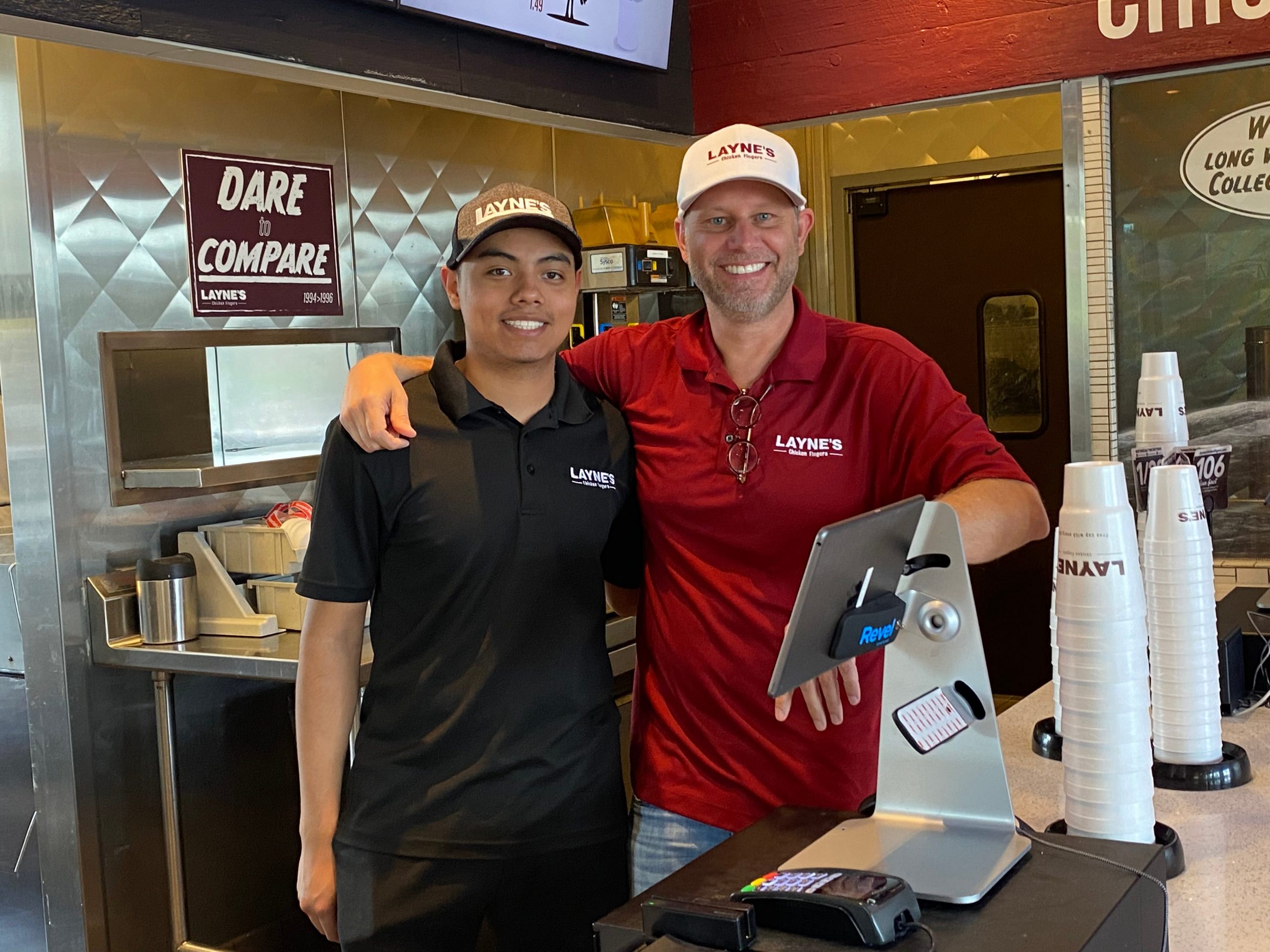 Two men stand behind the counter at a fast food restaurant smiling at the camera with their arms around each other.