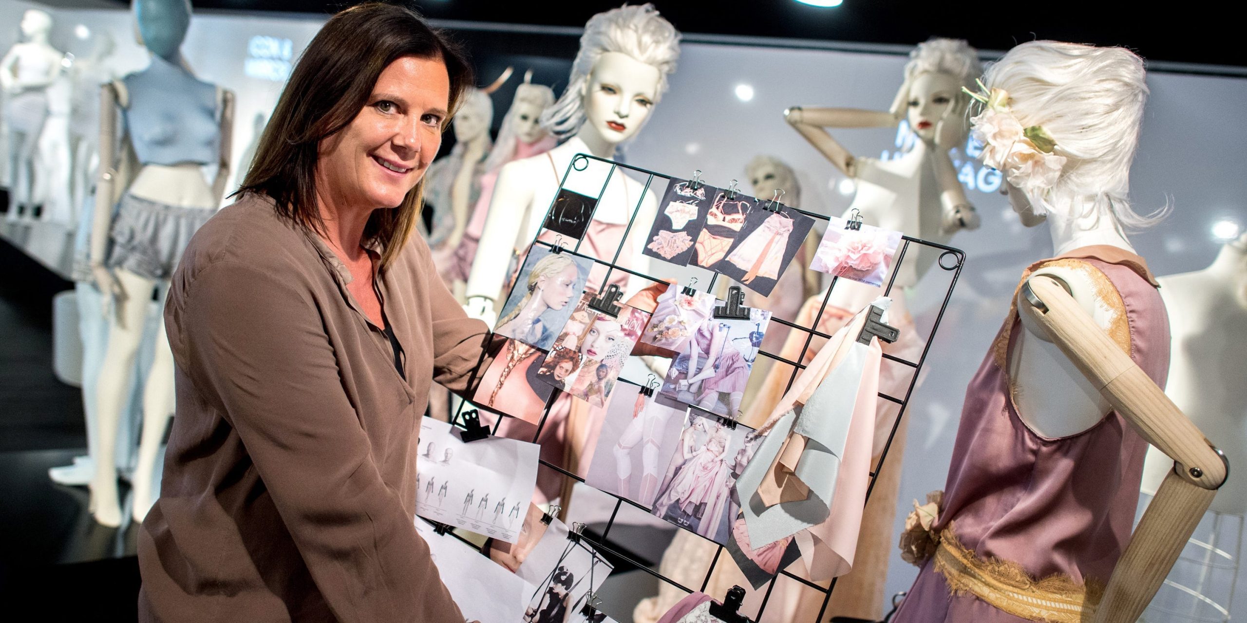 A woman holds a mood board filled with pictures of clothes and colors in front of some mannequins.