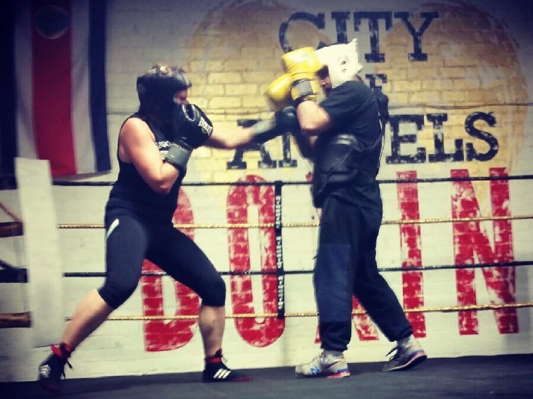 Two people wearing casual exercise clothing are seen sparring in a boxing ring.