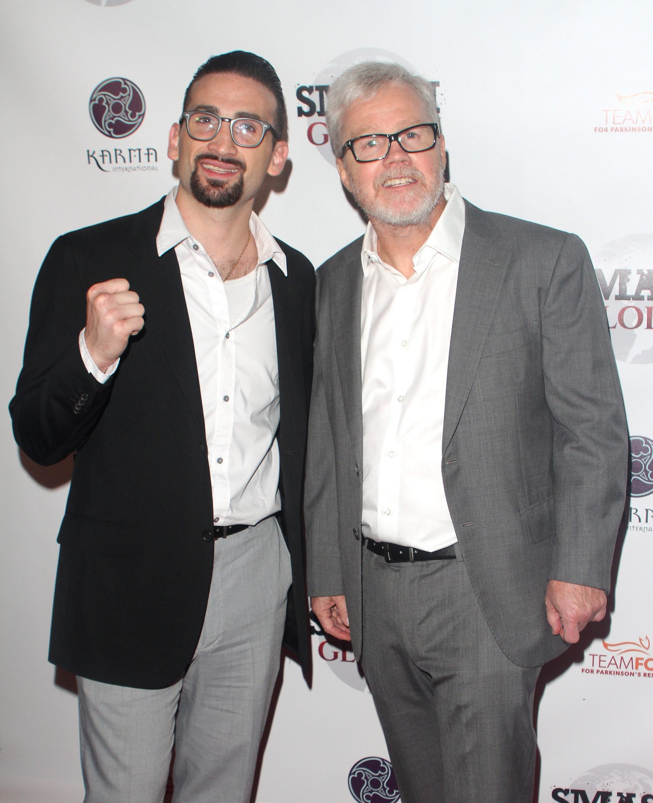 Two men wearing grey suits stand side by side, as one pumps his fist