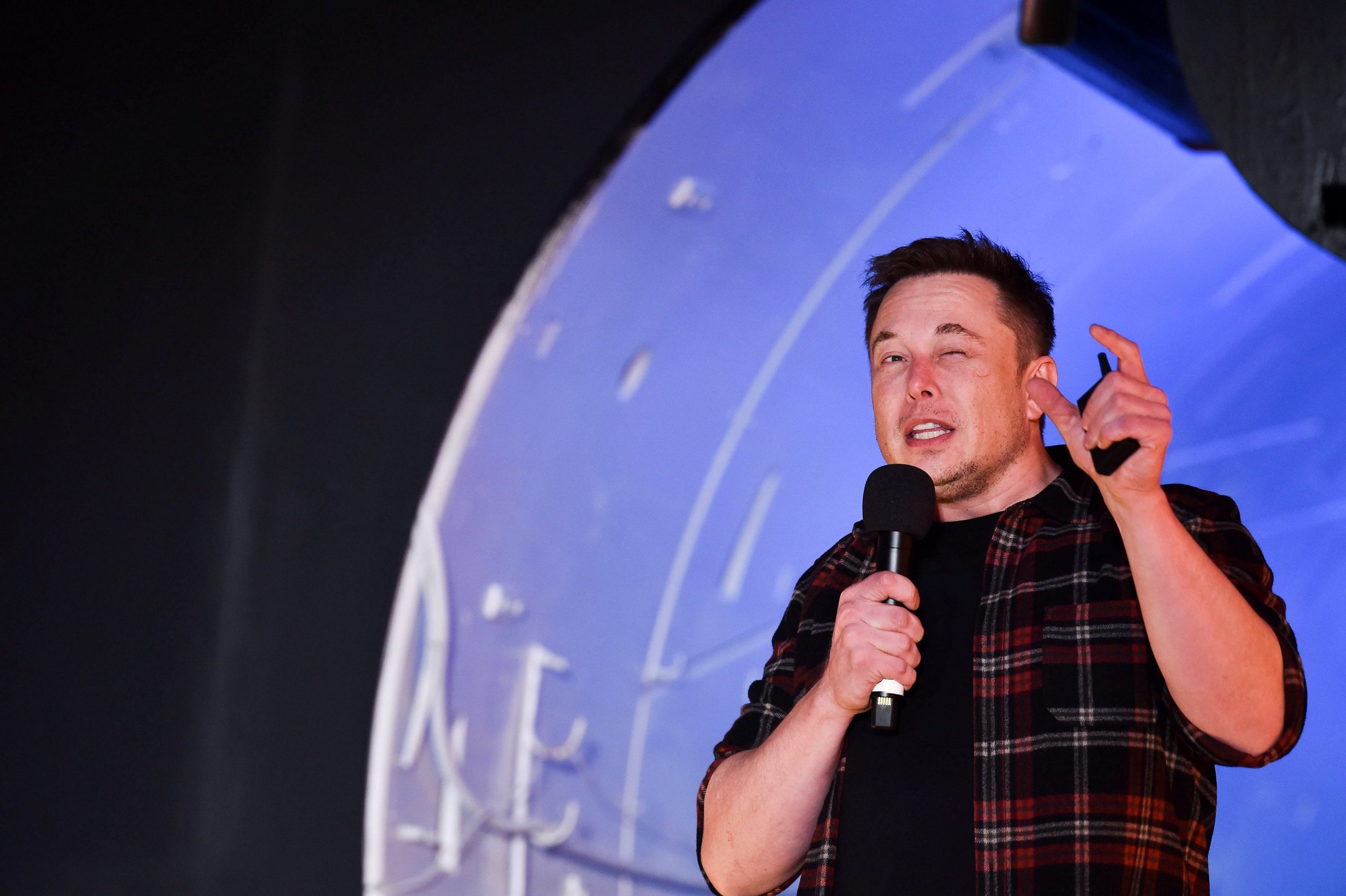 The Boring Company founder Elon Musk talks into a microphone in front of a tunnel lit in blue